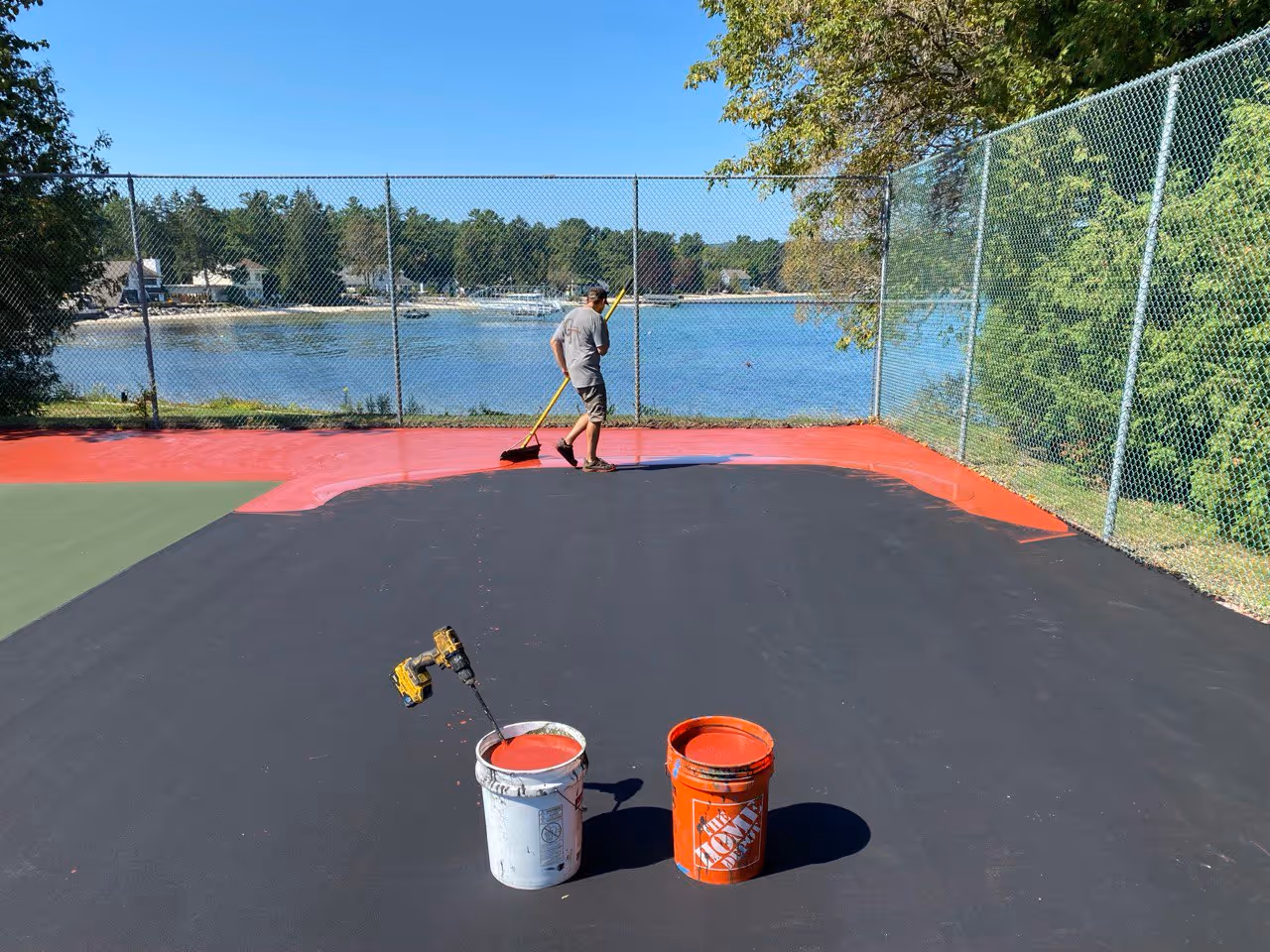 A professional crew member spreading the new vibrant red SportMaster color coat over a black acrylic resurfacer to define the court's outer border.