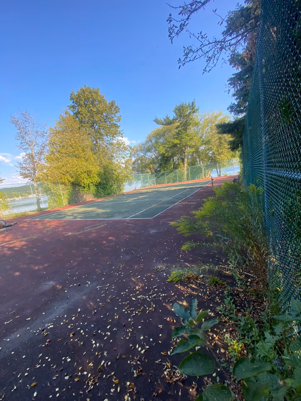 A wide view of the original dark red and green tennis court near Lake Michigan, featuring faded colors and weeds growing through the perimeter fence before restoration.