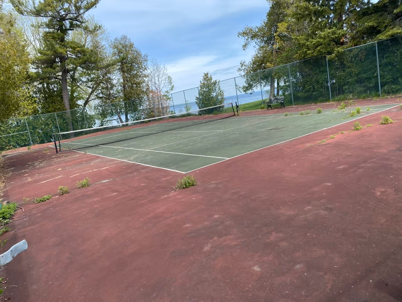 Initial "before" view of a deteriorated private tennis court on S Shore Dr in Suttons Bay, showing severe cracking and significant weed growth through the asphalt surface.