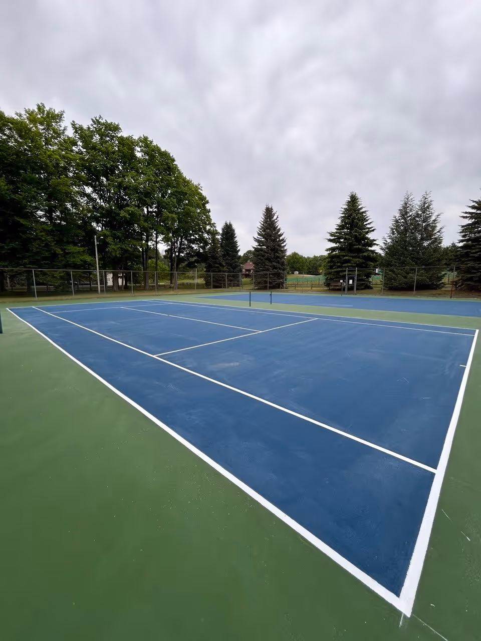 A wide view of the double tennis courts with crisp, new white lines painted. The high-contrast white SportMaster line paint looks vibrant against the deep blue and green acrylic surfacing.