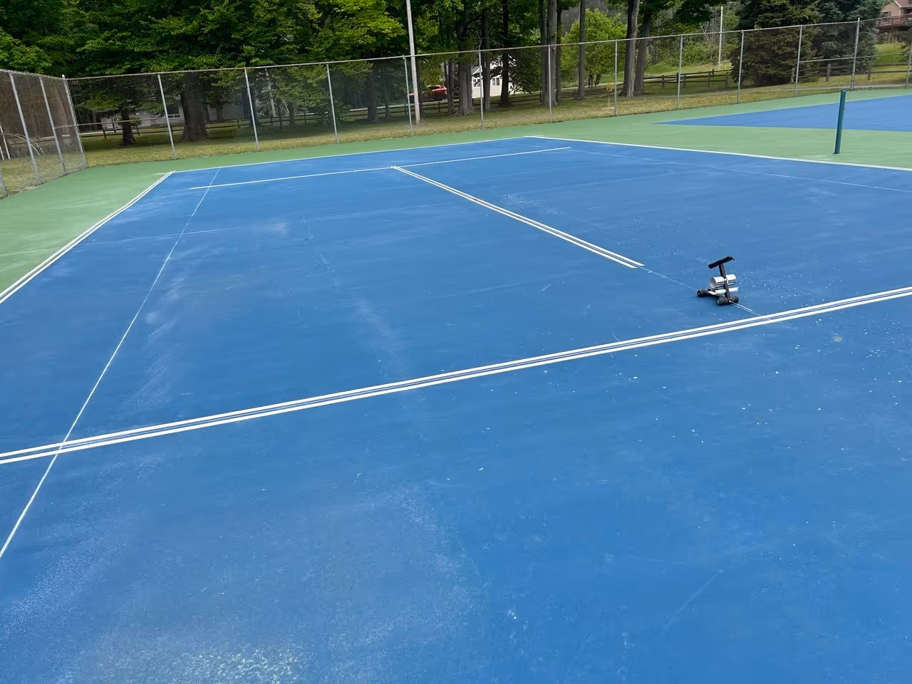 A specialized taping machine laying out the white lines for a tennis court. This technical photo highlights the meticulous precision required for professional tournament-spec striping on a large multi-court surface in Bowers Harbor Park.