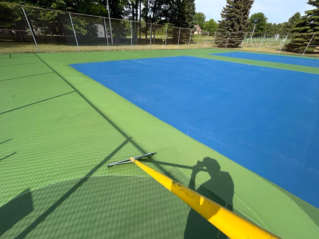An angled action shot showing a professional crew member spreading the vibrant green SportMaster color coat to define the outside boundary around the newly applied blue playing surface on the Old Mission Peninsula.
