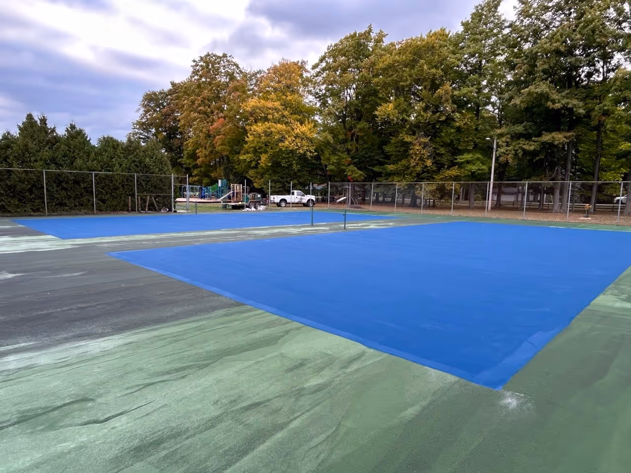 A progress shot of the double tennis courts in Traverse City. This view shows the extensive outside boundary area covered in black acrylic resurfacer, while the main internal play area is receiving its vibrant first SportMaster blue color coat.