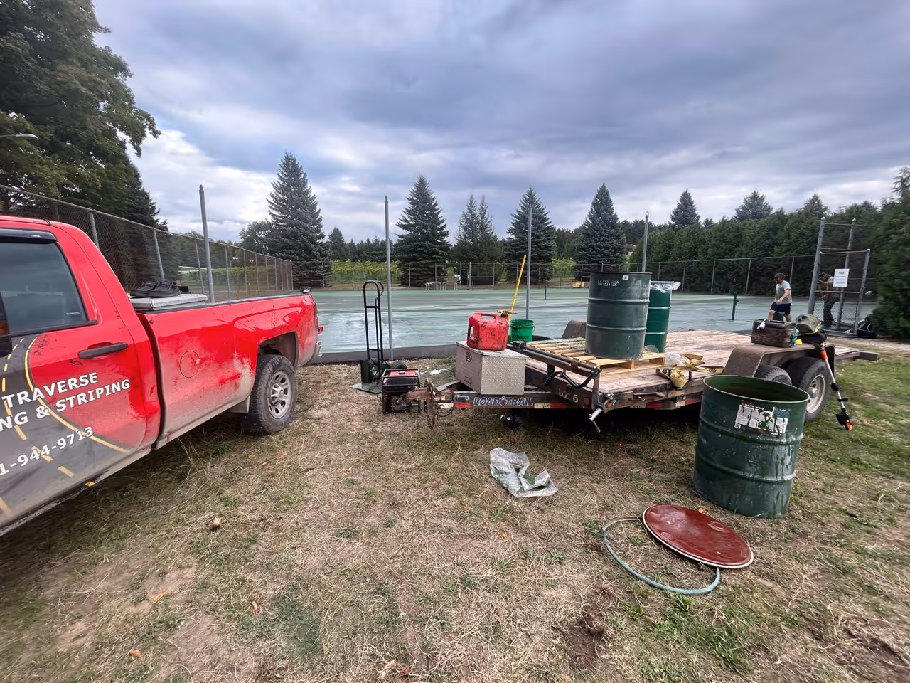 Professional mobilization at Bowers Harbor Park on the Old Mission Peninsula for a double tennis court resurfacing. The image shows the work truck, trailer, multiple 55-gallon drums of SportMaster materials, and the specialized mixing station on a flatbed trailer, with the existing courts in the background.