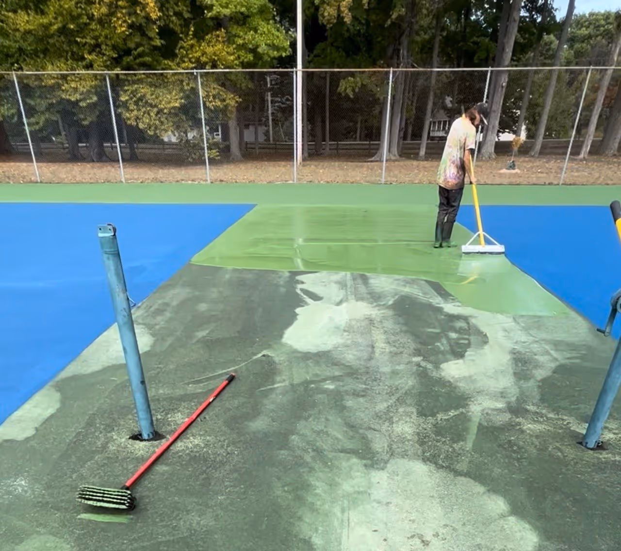 An action shot of a professional worker using a squeegee to spread the first coat of vibrant SportMaster green color coating on the space between the double tennis courts. The primary blue play areas are clearly visible on both sides, demonstrating the scale and technical precision of the large-scale resurfacing project at Bowers Harbor Park on the Old Mission Peninsula.