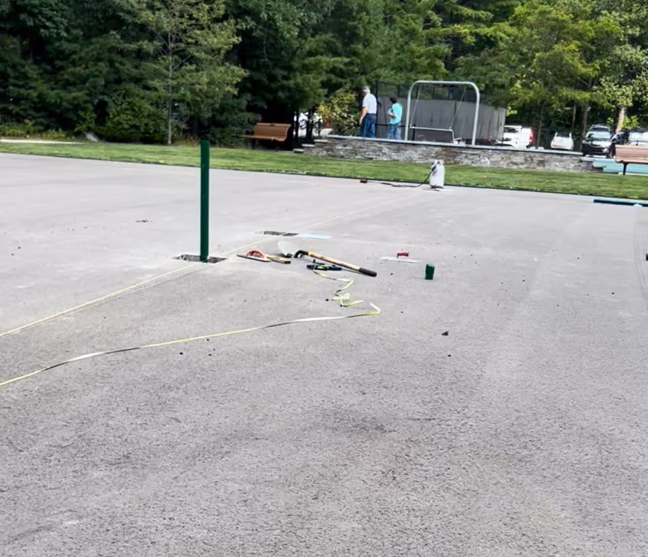 Professional tennis court net posts laid out on the asphalt during the installation phase, with one post successfully set in concrete.
