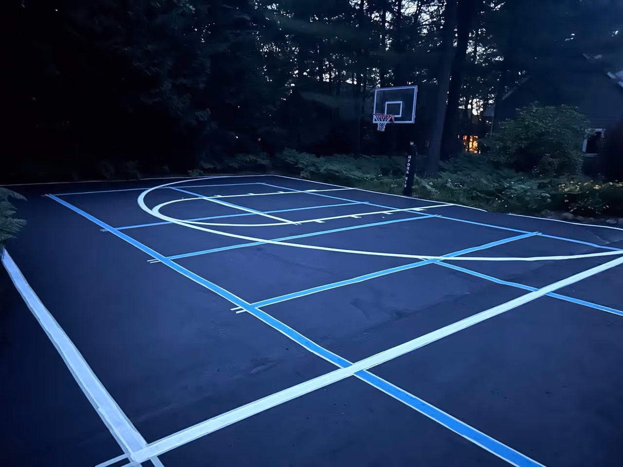 A "before" photo of a residential asphalt driveway on Long Lake in Traverse City, MI, showing a grey, faded surface with chipping blue pickleball lines and worn white basketball striping.