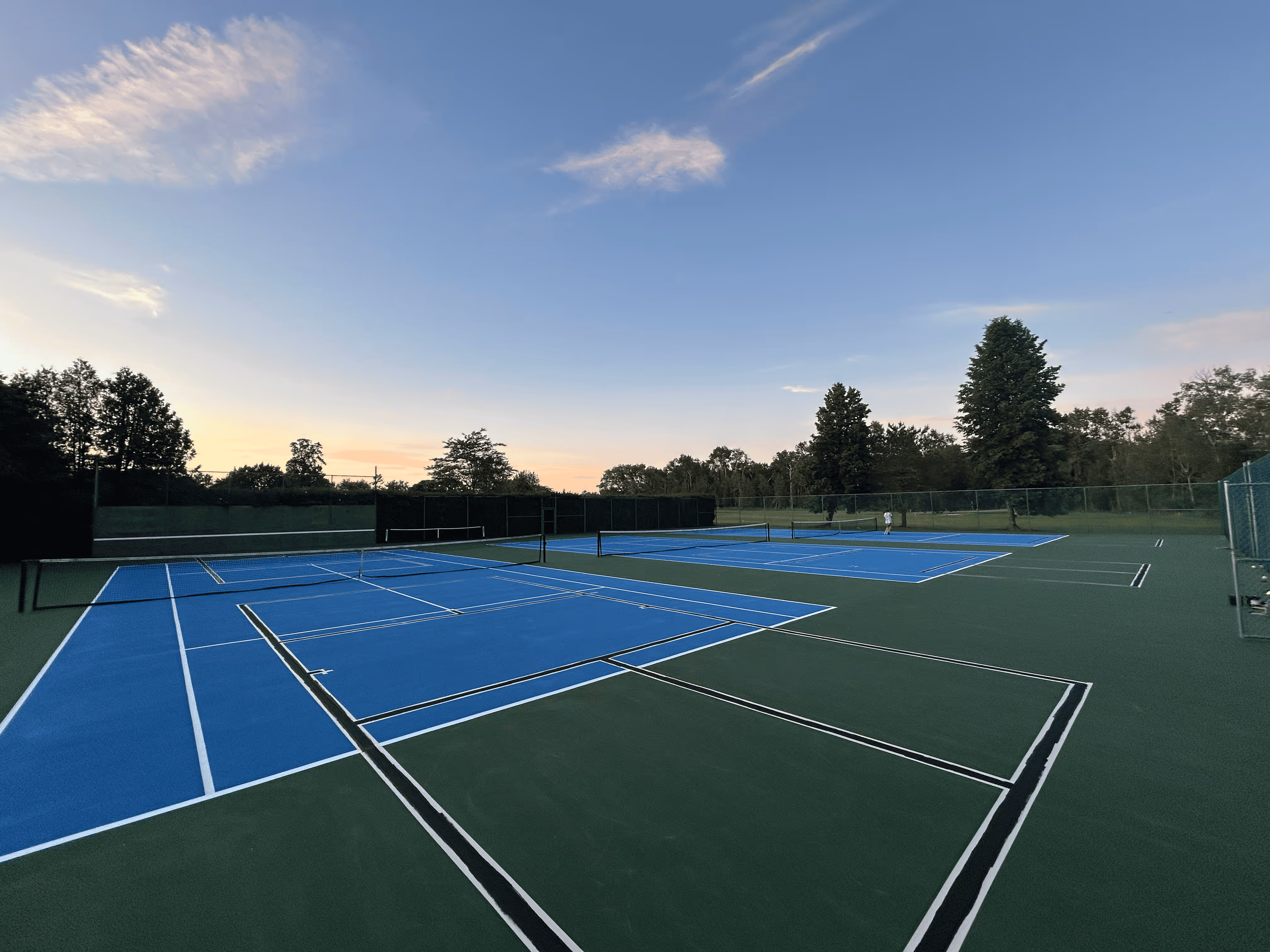 The completed triple tennis court complex at the Northport Point Homeowners Association on Northcott Dr. in Northport, MI. The vibrant blue and green SportMaster surface features precision white tennis lines and subtle black pickleball lines, accommodating six pickleball courts within the professional tennis facility.