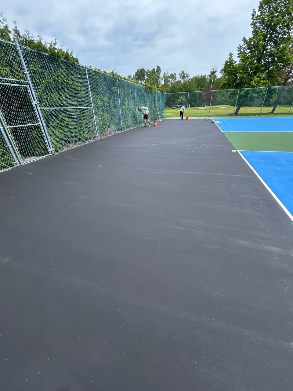 Professional crew members using squeegees to spread the first vibrant green SportMaster color coat around the perimeter boundary of the large triple-court play area in Northport.
