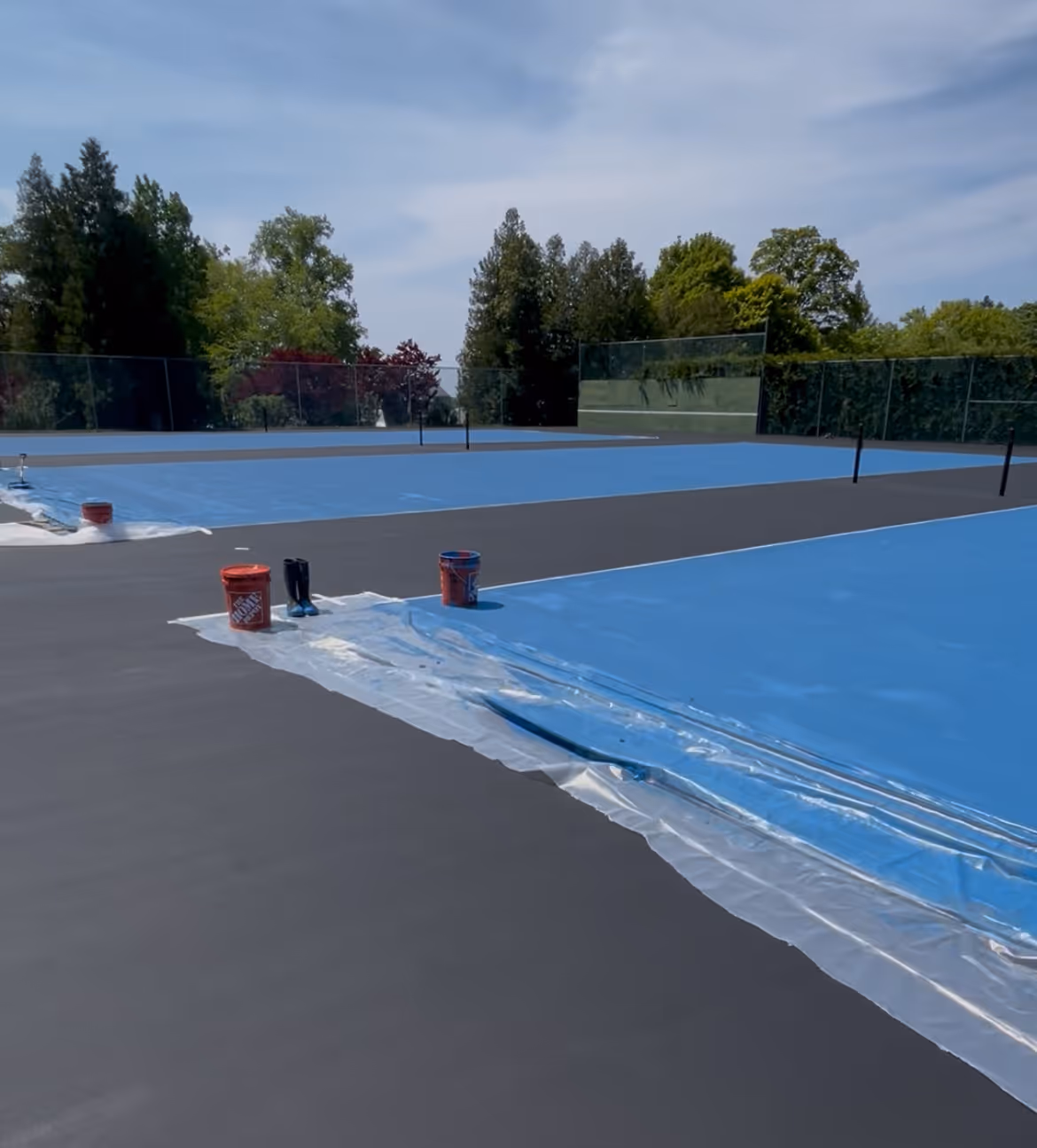A progress shot showing the black resurfacer boundary with all three primary tennis play areas successfully coated in vibrant SportMaster blue at the private Northport Point club.