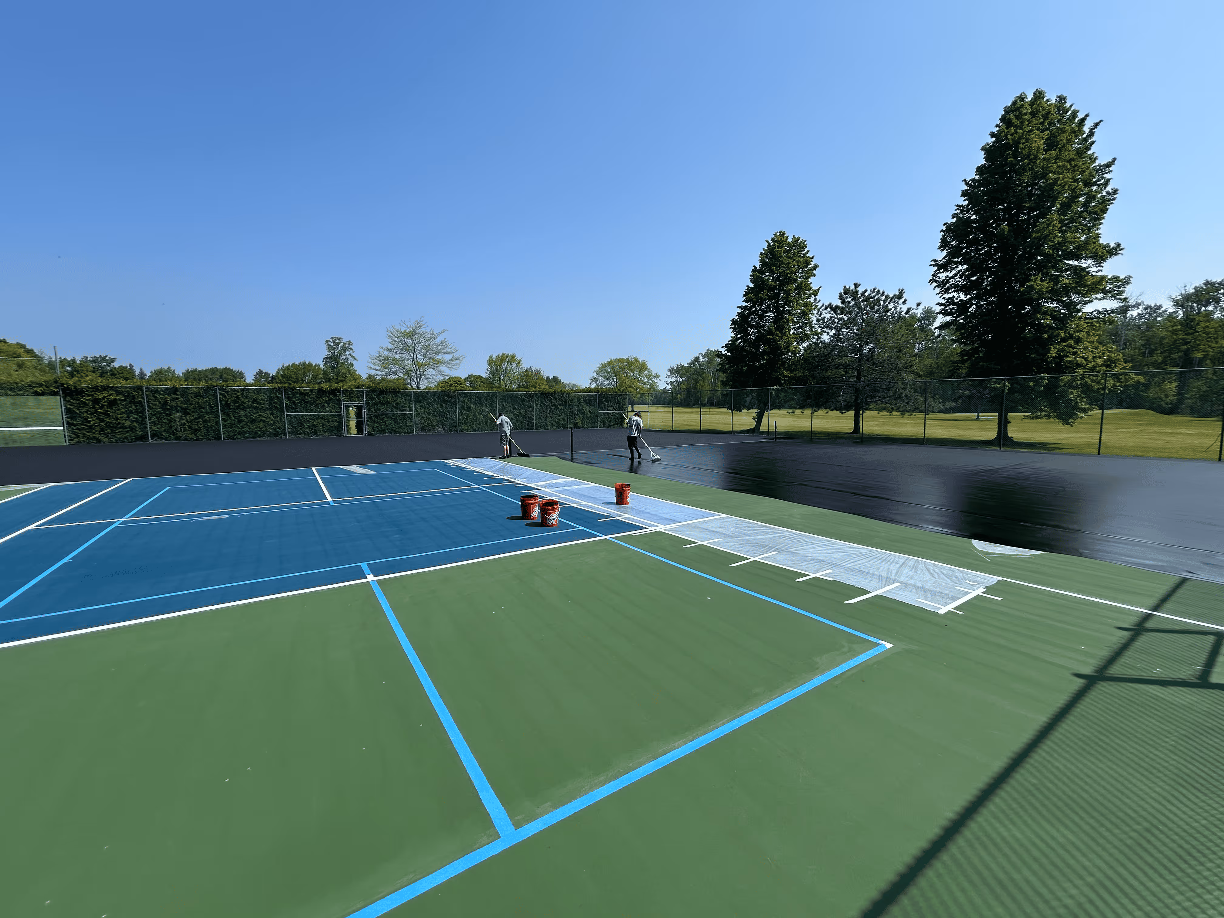 Two workers spreading the black acrylic resurfacer coat across the expansive asphalt surface, creating a smooth, uniform foundation for the multi-sport court system on Northcott Dr.