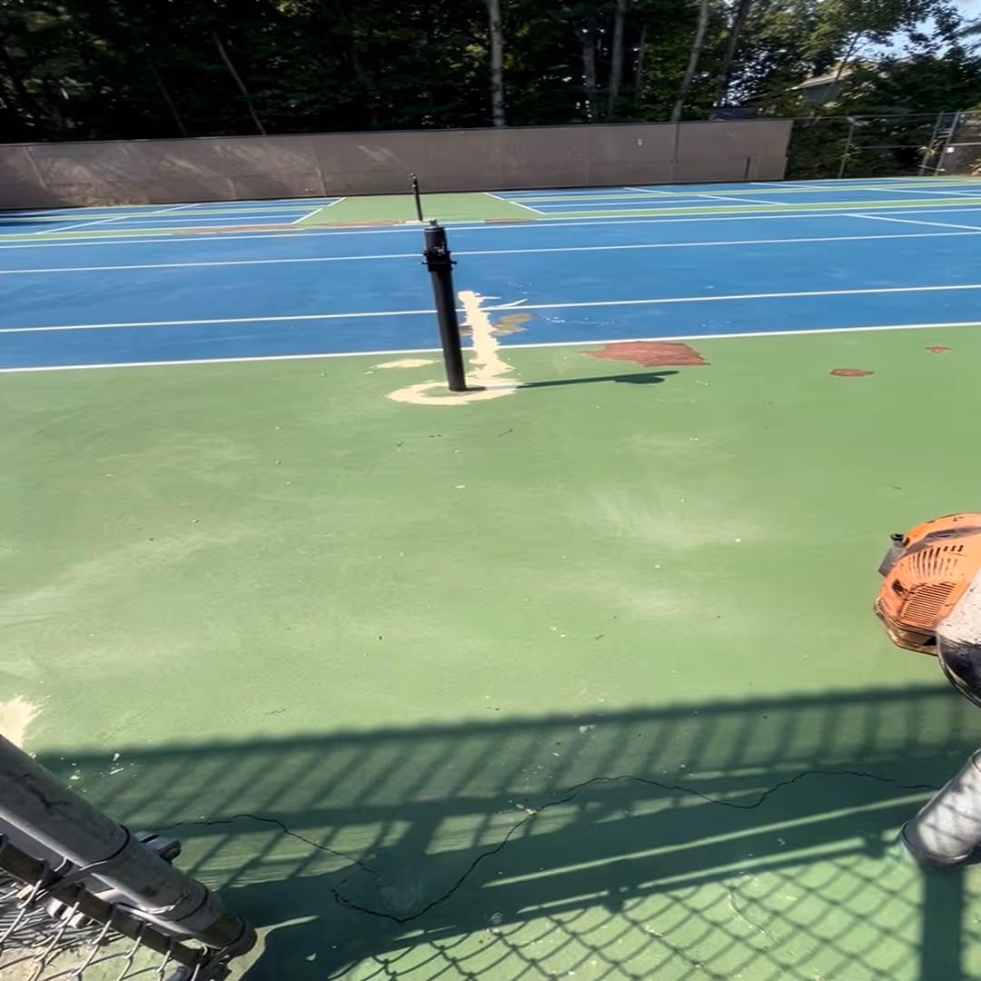 The beginning state of the single tennis court at Leland Public Schools before the project start. The image shows significant deterioration, with chunks of the surface missing (chipping) and existing cracks that have been filled, highlighting the need for a comprehensive professional resurfacing.