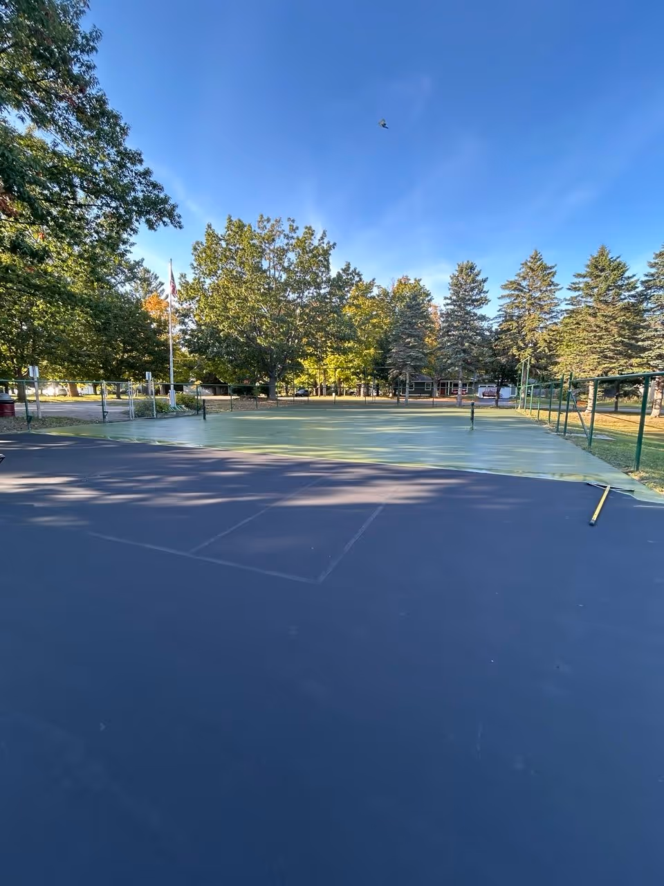 An action shot at Finch Park in Arcadia, MI, during a tennis court resurfacing project. The surface is split: half is covered in black acrylic resurfacer, while the first vibrant coat of SportMaster green color coat is being spread over the remaining half.