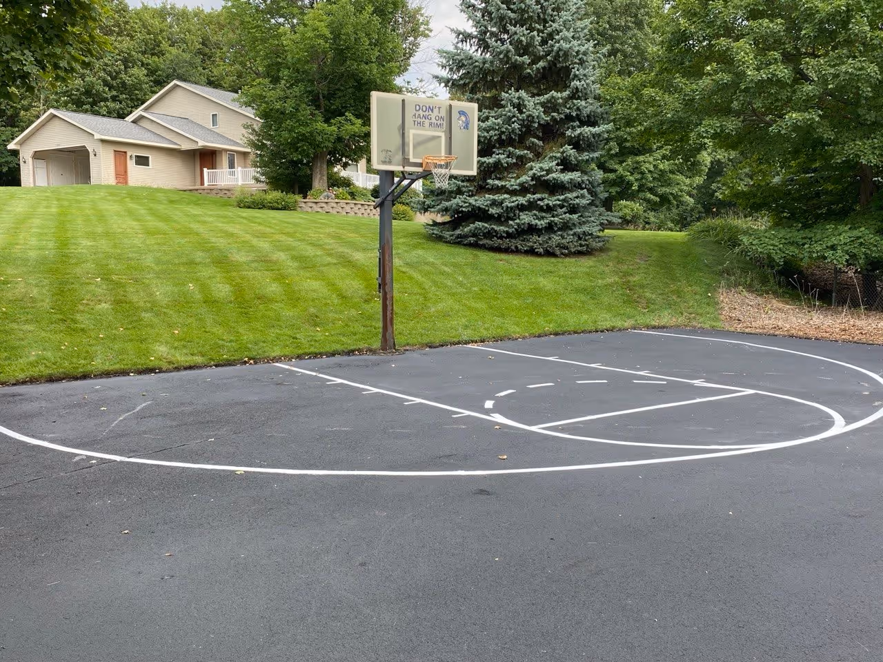 A view of a newly sealcoated residential asphalt driveway, showing a rich, jet-black finish. Precise white lines have been professionally striped to create a basketball court playing area directly on the fresh surface, including the free-throw line and key.