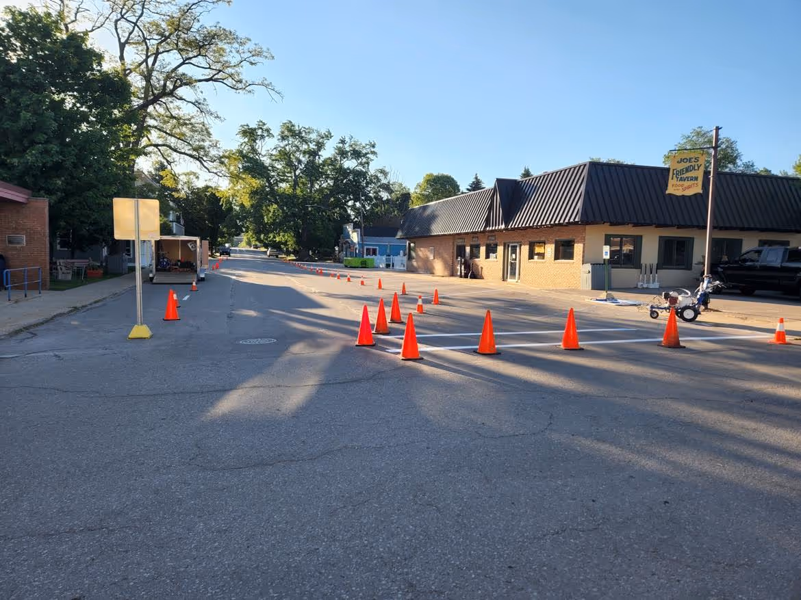 A professional road restriping and downtown parking project for the Village of Empire, MI. The image shows active pavement marking and traffic direction on Front St in front of Joe's Friendly Tavern, with safety cones and organized traffic control to ensure a smooth restoration of village parking and road lines.