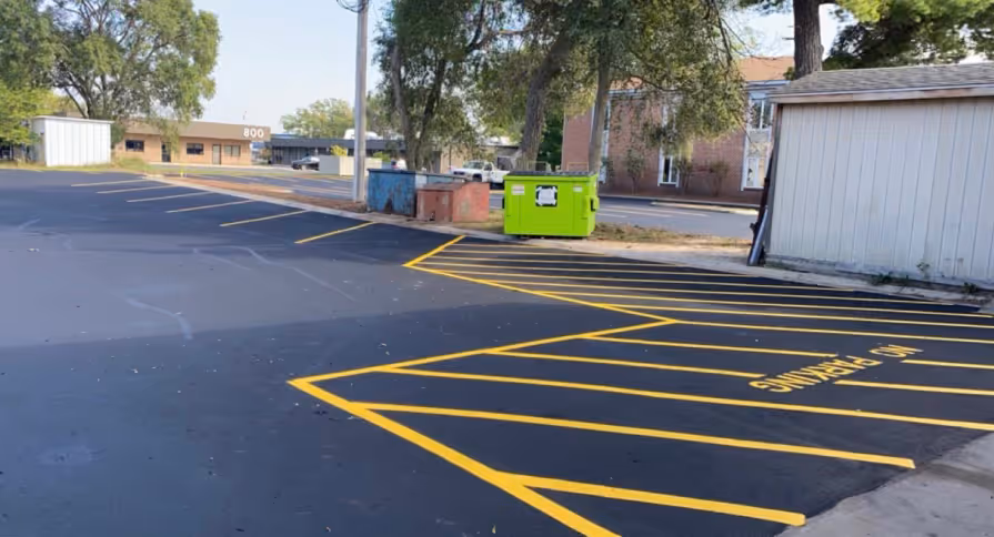 Precision parking lot striping and yellow cross-hatch no-parking zone markings at the Garfield Auto Service Center on S Garfield Ave in Traverse City, MI. The image showcases vibrant yellow pavement markings on a freshly sealed jet-black asphalt surface, providing clear traffic organization and safety for the busy auto repair facility.