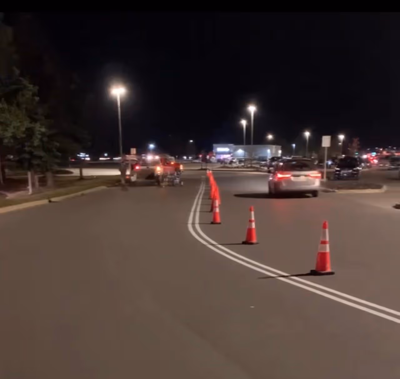 Professional road striping of curved double white lines at the Grand Traverse Mall off South Airport Rd in Traverse City, MI. The image shows active traffic control with safety cones, work trucks with flashing bubble lights, and a Graco 3900 LineLazer precision striper in operation on a high-traffic commercial perimeter road.