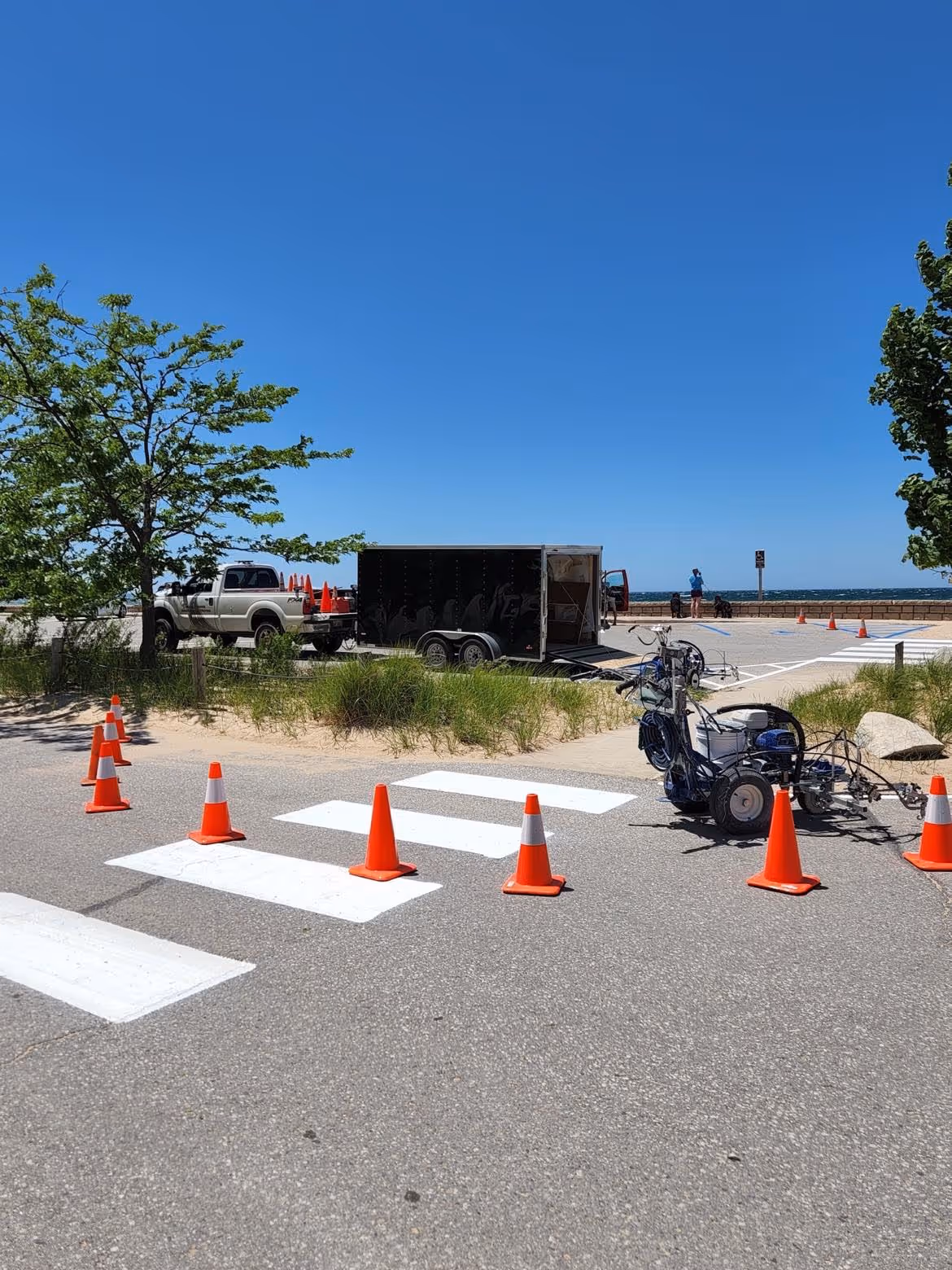 Active parking lot and crosswalk restriping in crisp white paint at the high-traffic Empire Beach public access for the Village of Empire, Michigan. The image shows orange traffic cones directing vehicle flow on Lake Michigan Dr and Niagara St, with a professional Graco LineLazer 3900 in use to ensure precision pavement markings near the Lake Michigan shoreline.