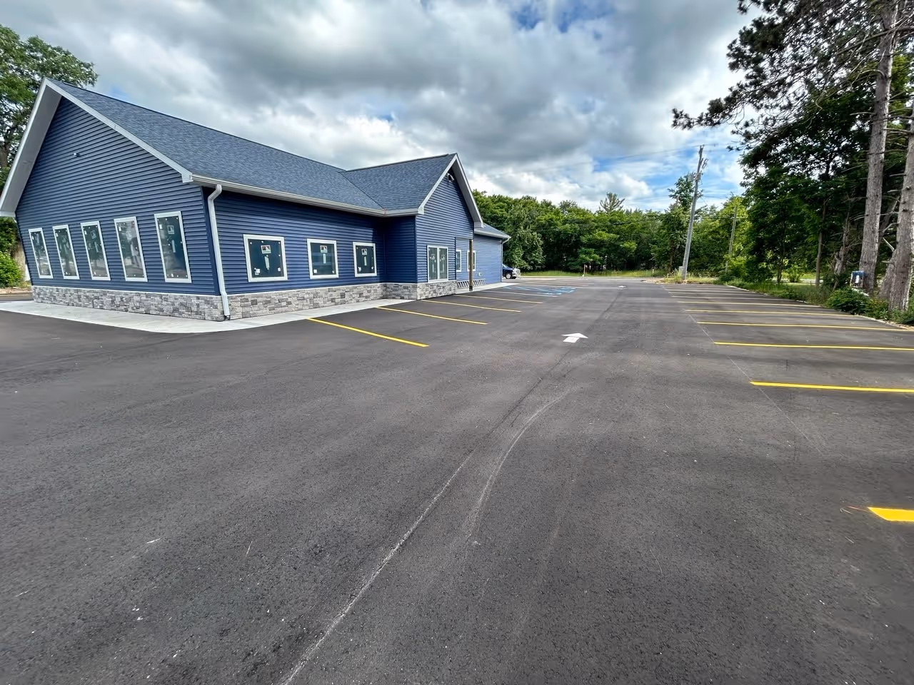 A freshly paved asphalt parking lot at B.C. Pizza in Bellaire, MI, featuring a complete new layout for Team Elmer's. The image showcases high-contrast yellow parking stall lines, blue ADA-compliant handicap stalls with symbols, and white directional arrows for optimized traffic flow and customer safety.