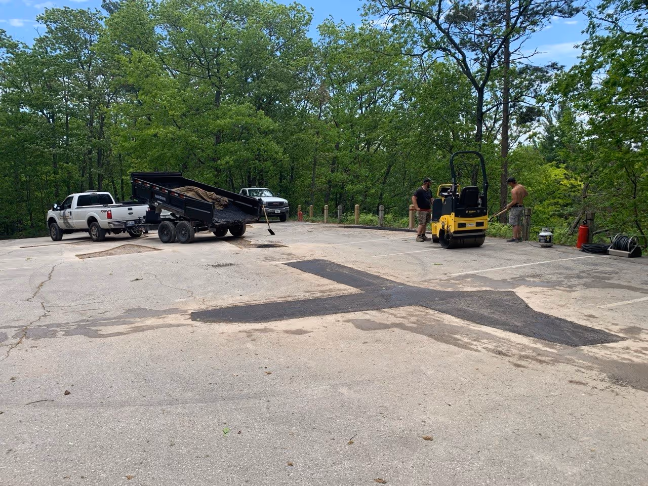 Professional asphalt crew preparing for a Cut & Replace repair at The Homestead Resort's Pinnacle Place Condo Association off Michigan Scenic Byway M-22 near Glen Arbor. The upper parking area shows one completed patch, two workers prepping the next section, a Bomag yellow asphalt roller, a Load Trail dump trailer, and a Ford F-350 truck.