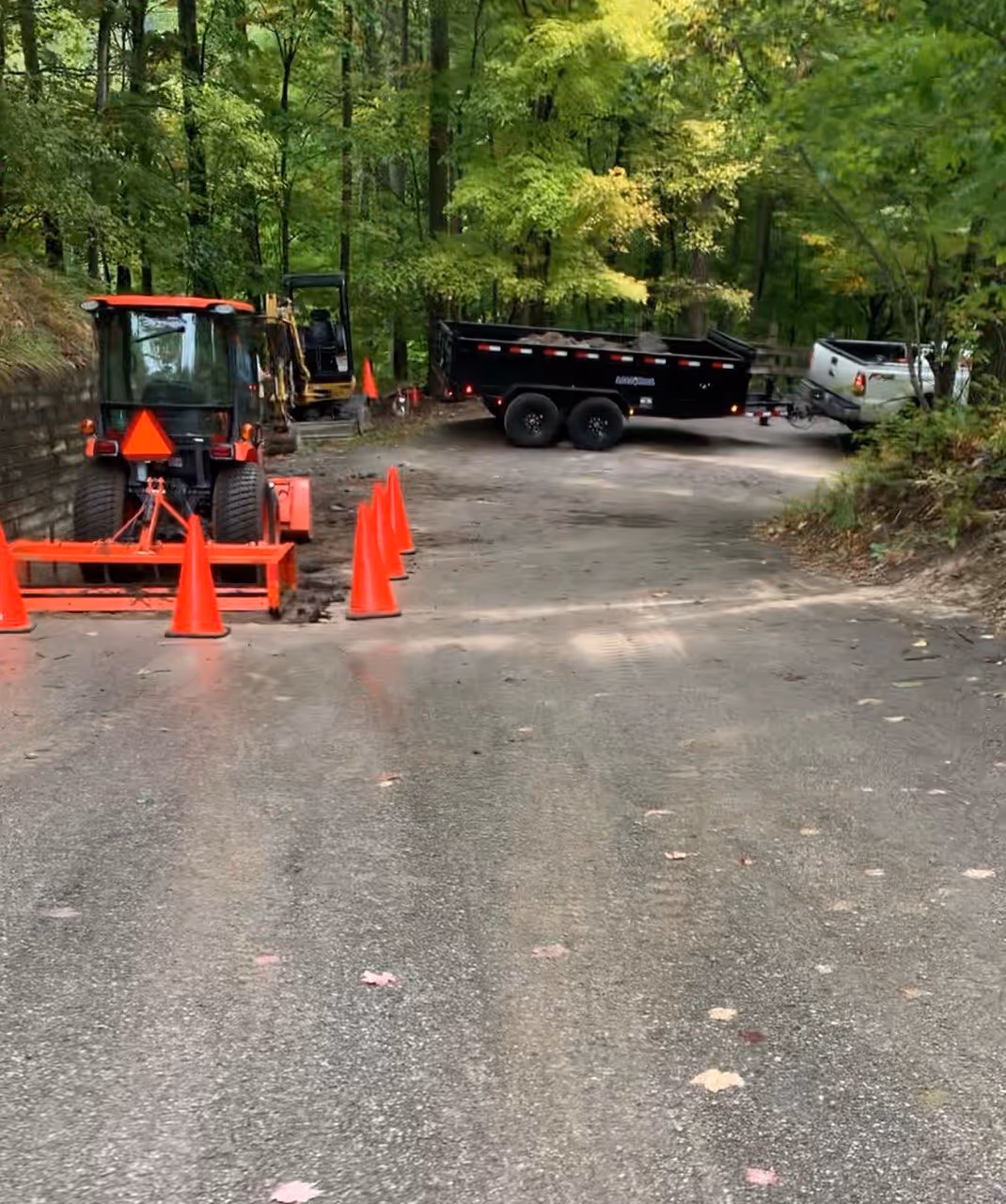 Active asphalt removal underway at The Homestead Resort off M-22 in Glen Arbor, MI. The work area is coned off with orange cones for safety while a Kubota tractor and a Caterpillar mini-excavator load a Load Trail dump trailer, assisted by a Ford F-350 truck for the Cut & Replace process.