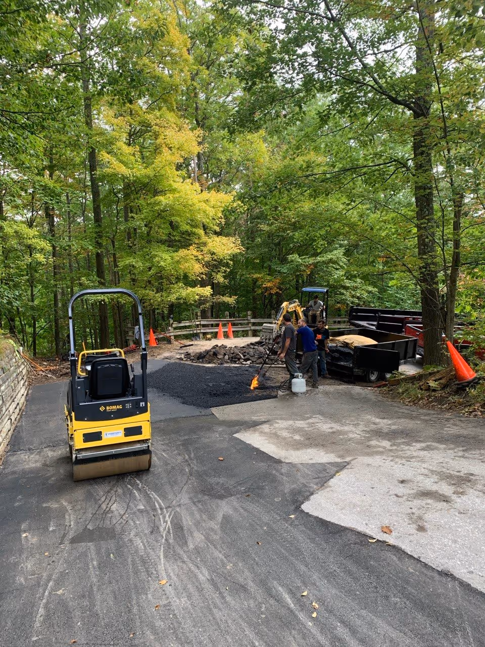 Asphalt workers torching and raking hot asphalt mix on a sharp corner at The Homestead Resort near Glen Arbor, MI. A Bomag yellow roller sits in wait, and a Caterpillar mini-excavator is simultaneously removing old asphalt for the adjacent patch area.