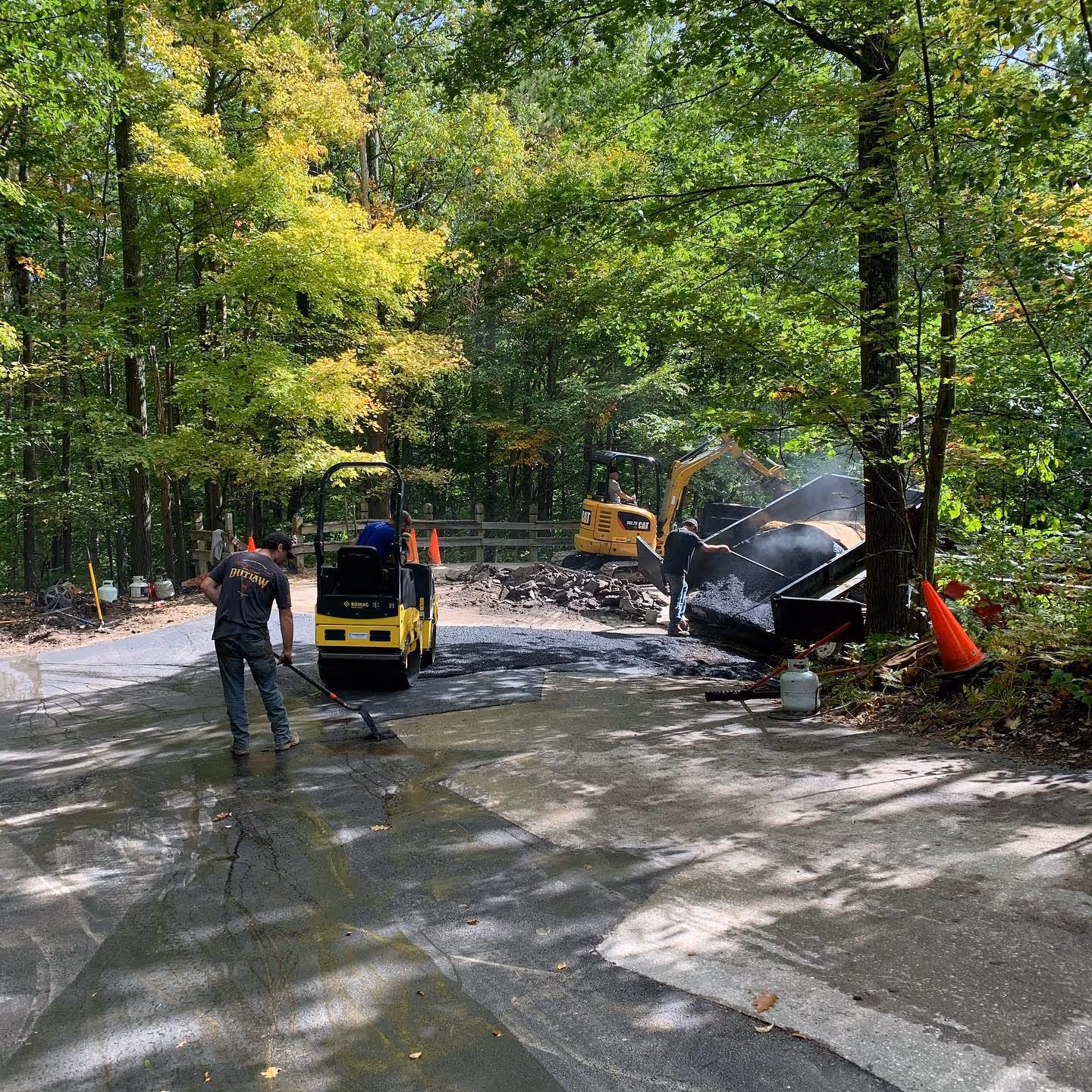 active asphalt repair work at The Homestead Resort off M-22. The image shows a Bomag roller compacting new asphalt, a worker shoveling steaming hot mix from a raised dump trailer, and a Caterpillar mini-excavator loading a second dump trailer with old pavement debris.