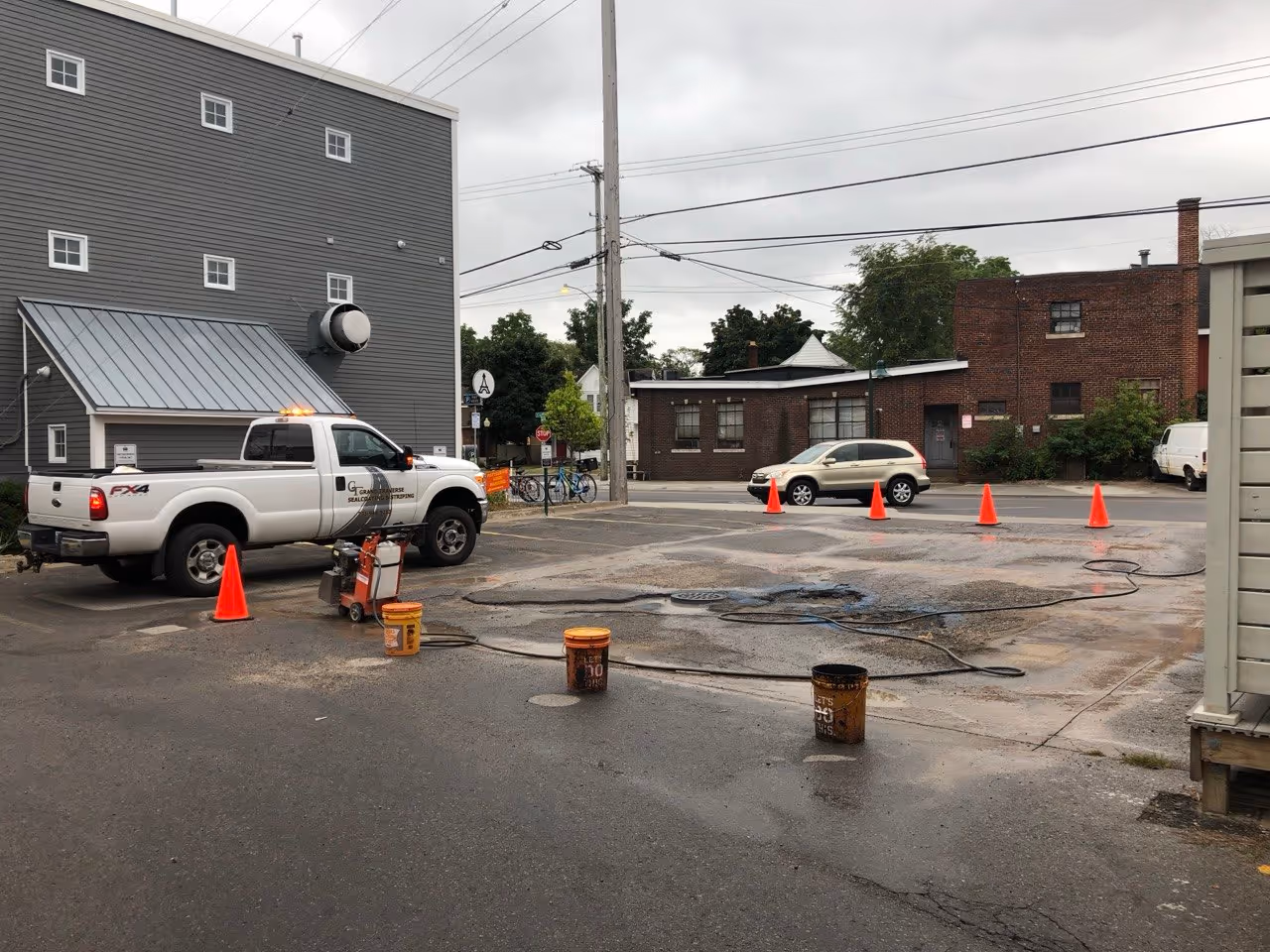 A self-propelled asphalt saw with water-cooling dust suppression cutting a repair perimeter at Rare Bird Brewpub in Traverse City, MI. A Grand Traverse Sealcoating & Striping Ford F-350 with safety lights secures the coned-off work zone on Lake Ave