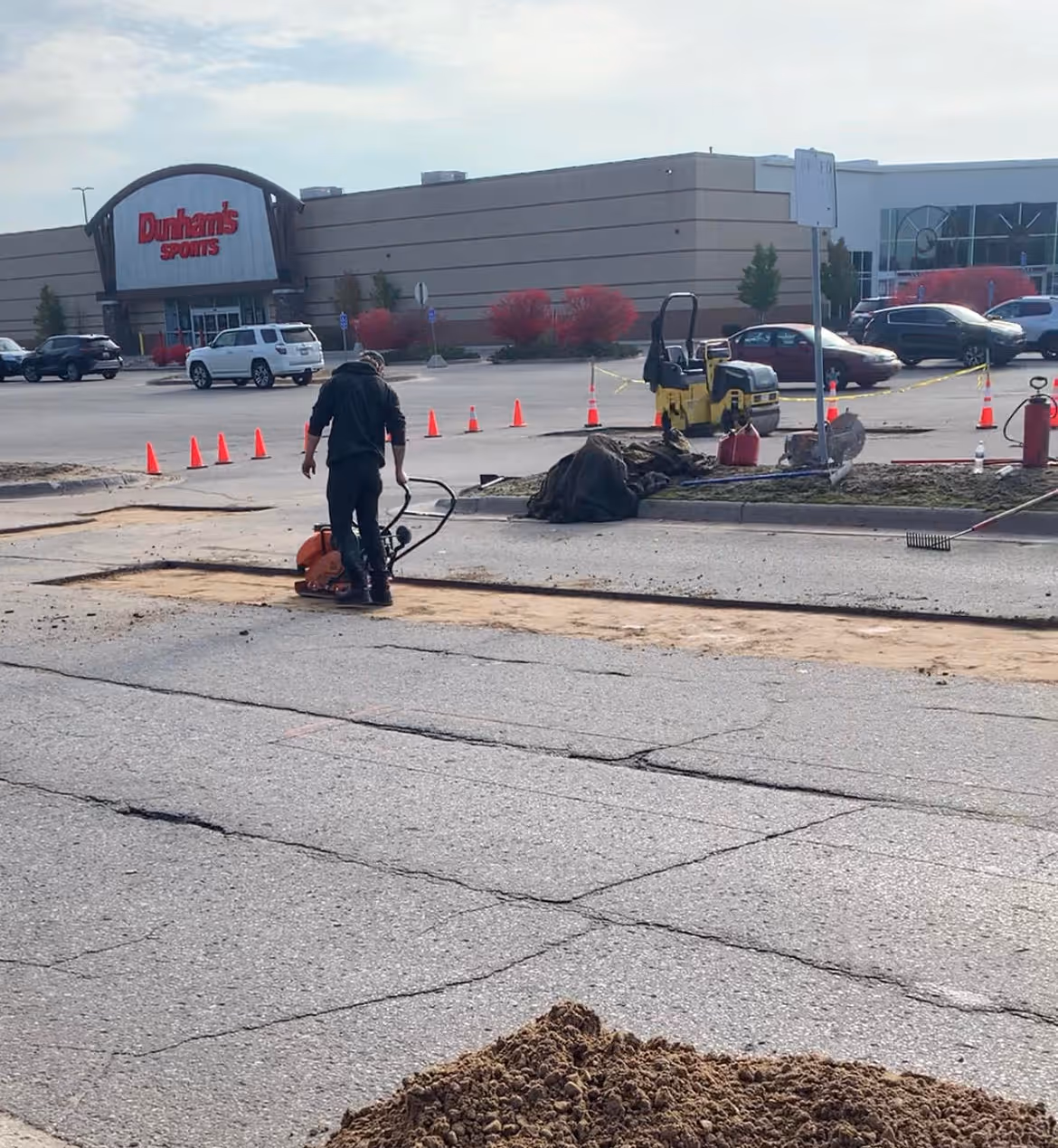 An asphalt worker using a precision plate compactor to stabilize a new gravel base at the Grand Traverse Mall road in Traverse City, MI. In the background, a Bomag roller and a second worker raking hot-mix asphalt show active structural road repair in front of Dunham’s Sports.