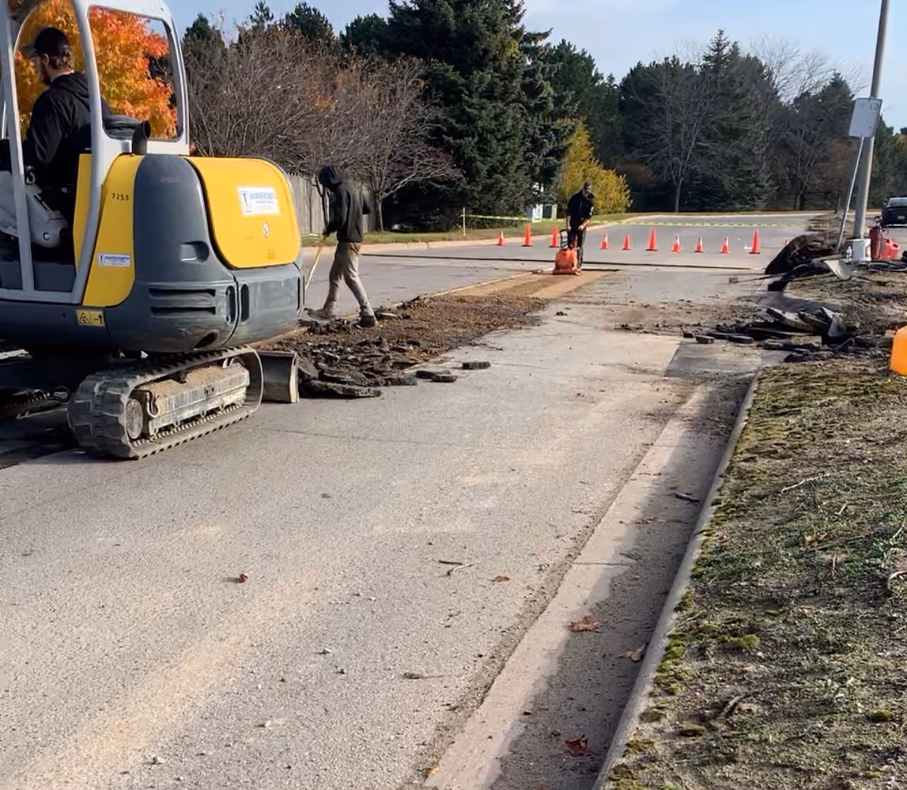 A Wacker Neuson 3507 excavator removing failed road asphalt on the Grand Traverse Mall perimeter road. The image shows a worker running a plate compactor in the background with orange traffic cones directing vehicle flow between US-31 and South Airport Rd.