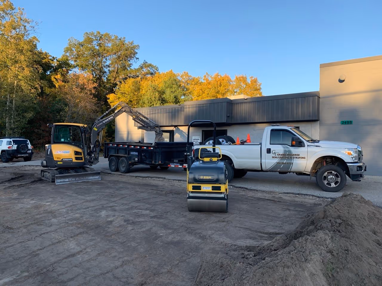 Site preparation for a parking lot extension at Threads Custom Gear on Cass Rd in Traverse City, MI. A Volvo ECR40D mini excavator loads old asphalt and topsoil into a Load Trail dump trailer, while a Bomag BW120 roller compacts new 22A road gravel to create a stable base.