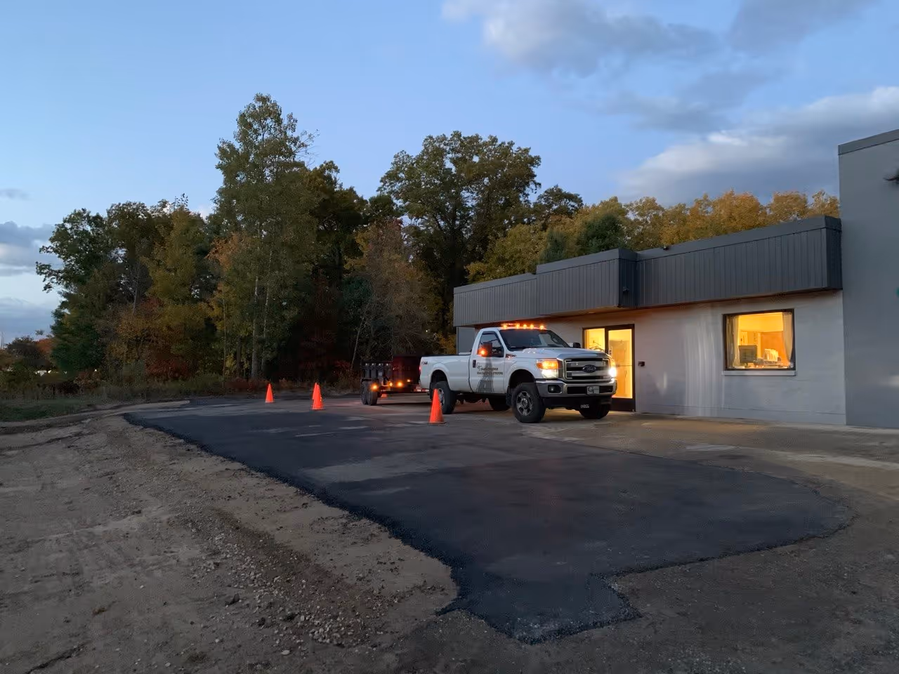 Freshly installed and compacted asphalt at the entrance of Threads Custom Gear in Traverse City. The area is protected by orange safety cones, with a Ford F250 and Load Trail dump trailer positioned in the background of the newly paved commercial lot.