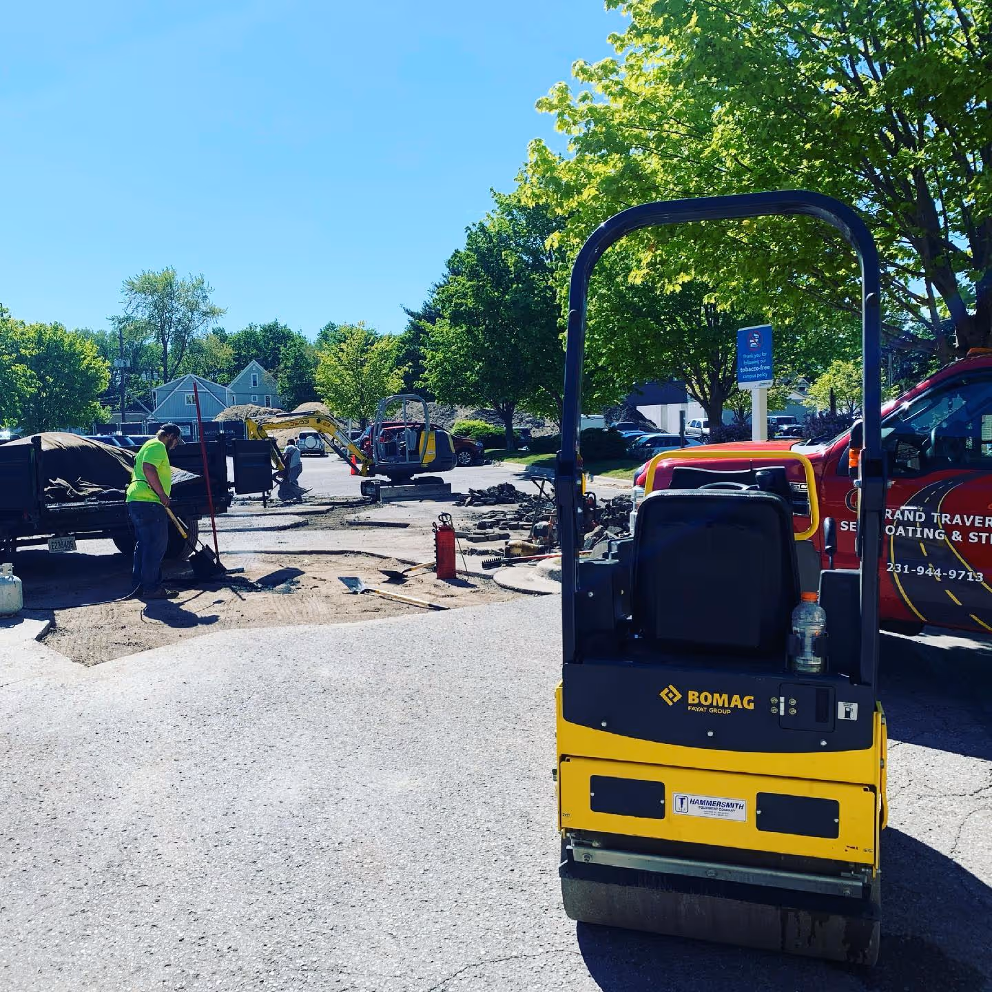 Comprehensive asphalt repair at a multi-company building in Traverse City managed by Schmidt-Rogers Management. A worker shovels new asphalt from a Load Trail dump trailer while a Wacker Neuson 3507 excavator and Bomag BW120 roller prep adjacent patches near the North Peak Brewing Company entrance.