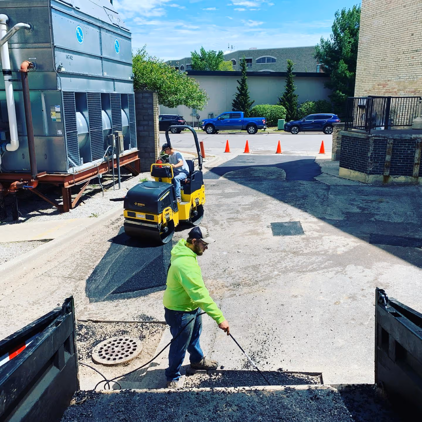 Professional asphalt patching for a complex housing Stafford-Smith and Kilkenny's. One worker operates a Bomag BW120 roller on a luted patch while another uses a torch to maintain asphalt temperature for a smooth finish. Orange cones secure the lot entrance near a prepped drainage repair.