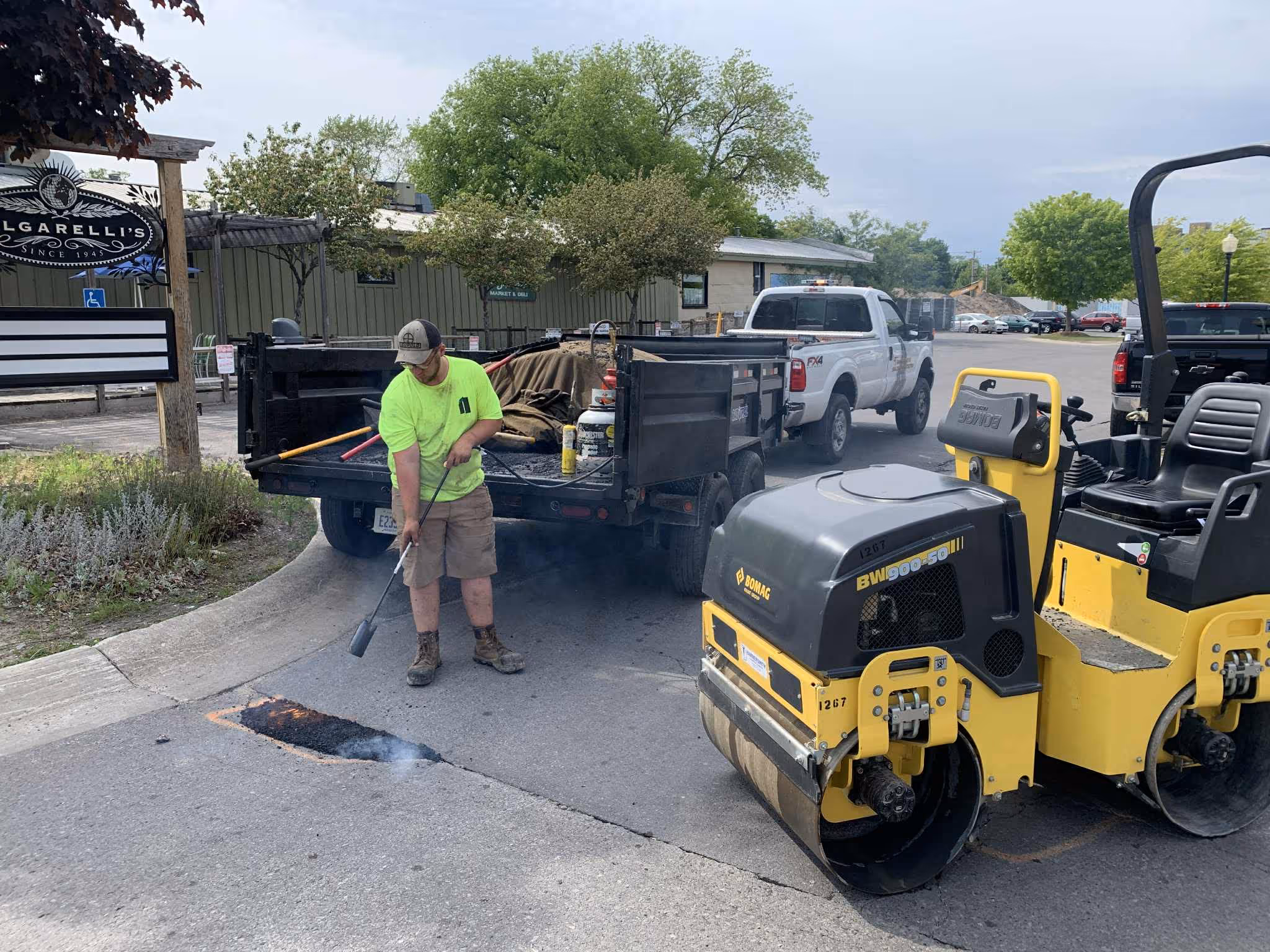Asphalt pothole repair at the shared entrance for Folgarelli's Market & Wine Shop in Traverse City. A worker uses a torch to heat edges for a thermal bond with new asphalt, supported by a Ford F-250 and a Load Trail dump trailer holding hot-mix under canvas tarps.
