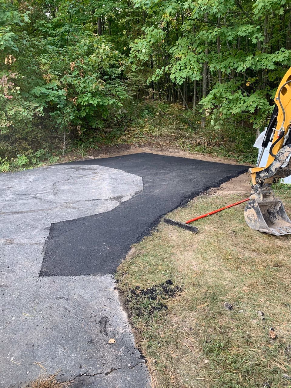 A close-up of a newly installed and smooth-compacted asphalt patch on a residential driveway. The image highlights the transition from the excavator bucket to the finished, level surface on this M-22 property.