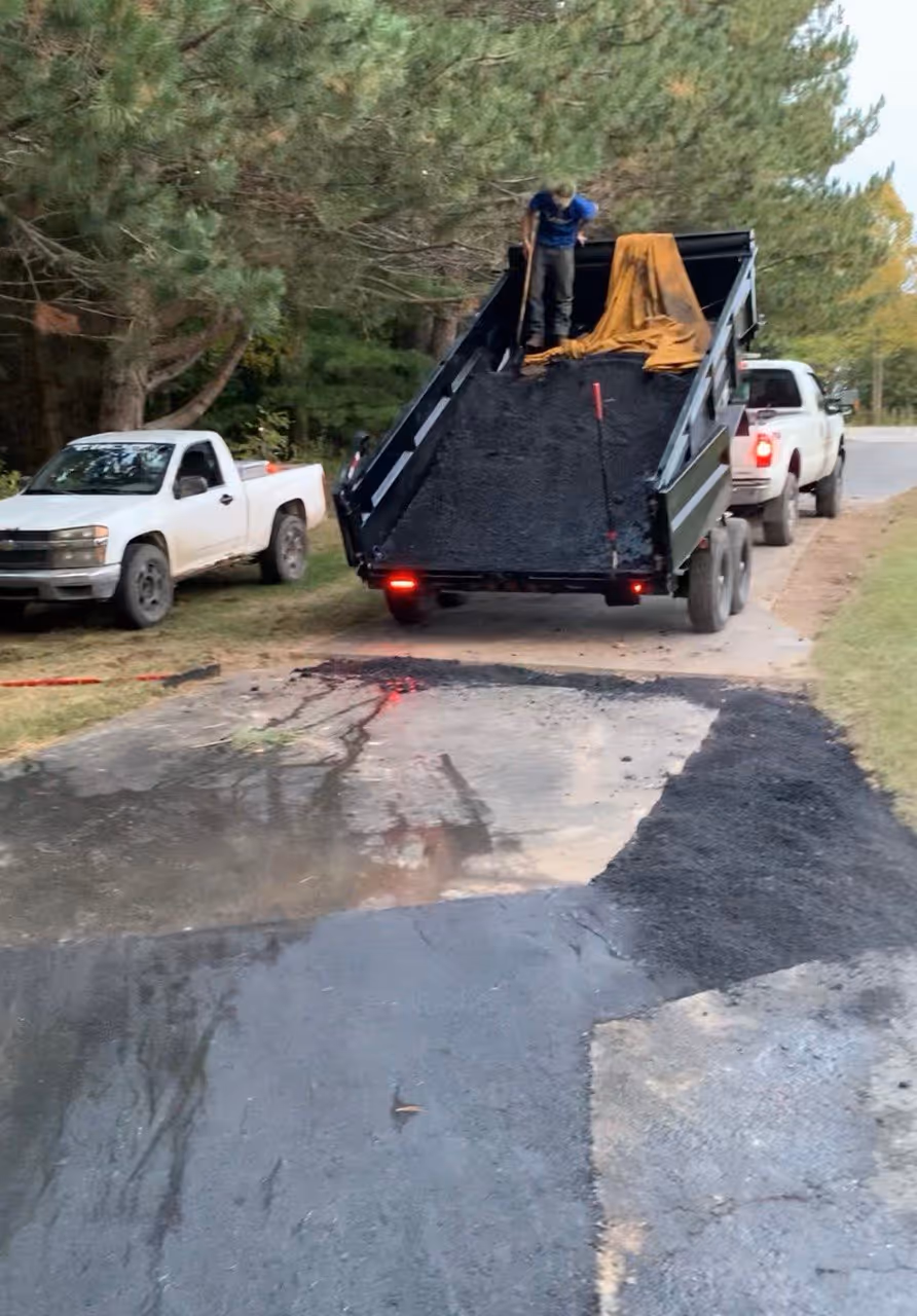 Active hot-mix asphalt installation on a Suttons Bay driveway. A worker shovels steaming asphalt from a raised Load Trail dump trailer into a prepped area, showing the transition from unraked material to a professional-grade patch.