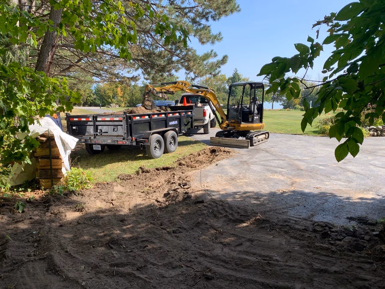 A Caterpillar 302.7D mini excavator removing failed asphalt and sub-base material from a driveway in Suttons Bay. The debris is loaded into a Load Trail dump trailer hooked to a Ford F-250 for efficient site clearing along the Pure Michigan Byway.