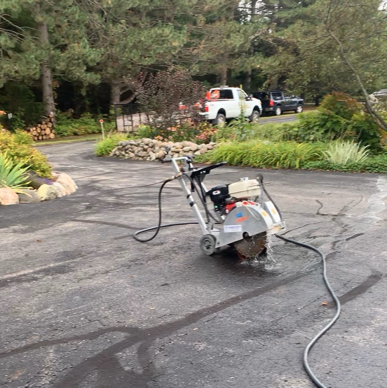 Initial stage of a residential driveway repair in Suttons Bay, MI on N West Bayshore Dr (M-22). A self-propelled asphalt saw with an active water-cooling system is shown cutting a precise repair perimeter while suppressing dust near Omena Cut Flowers.