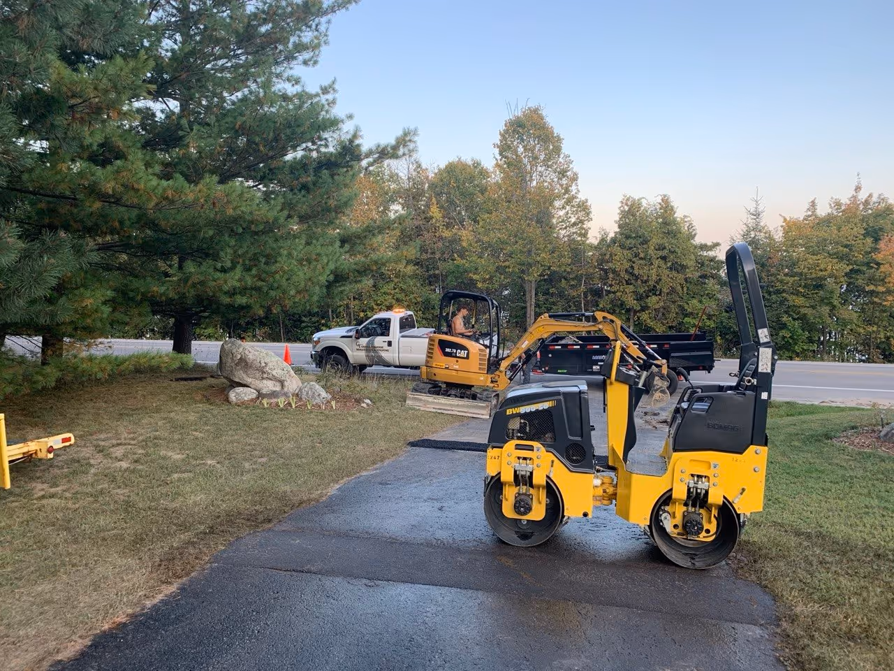 A systematic repair sequence moving toward the M-22 entrance. A Bomag BW120 roller compacts a fresh patch while the Caterpillar 302.7D removes damaged asphalt in the background, ensuring the mix stays hot and workable throughout the driveway.