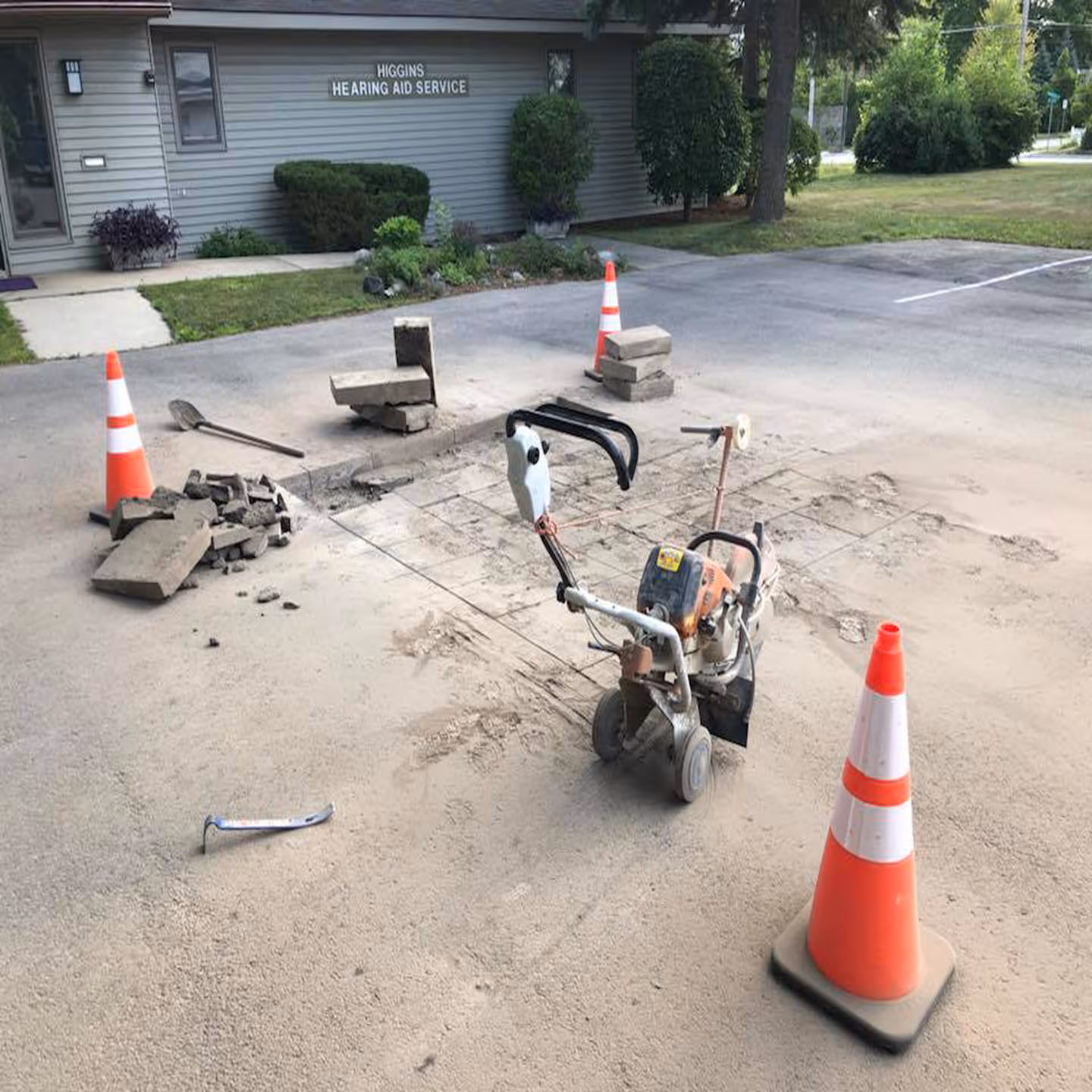 Initial phase of an asphalt repair at Higgins Hearing and Audiology on W Front St in Traverse City, MI. The image shows a coned-off work zone with a Stihl TS800 saw on a cart, with the damaged asphalt precisely cut into sections for structural removal.