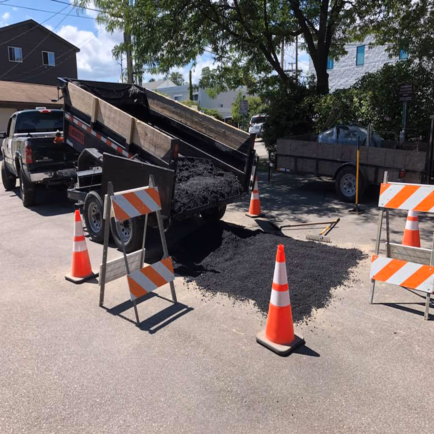 Active hot-mix asphalt installation at the Higgins Hearing parking lot. The repair area is half-filled with steaming asphalt while a Chevy 2500 Duramax Diesel provides support with a raised dump trailer delivering fresh, workable material.