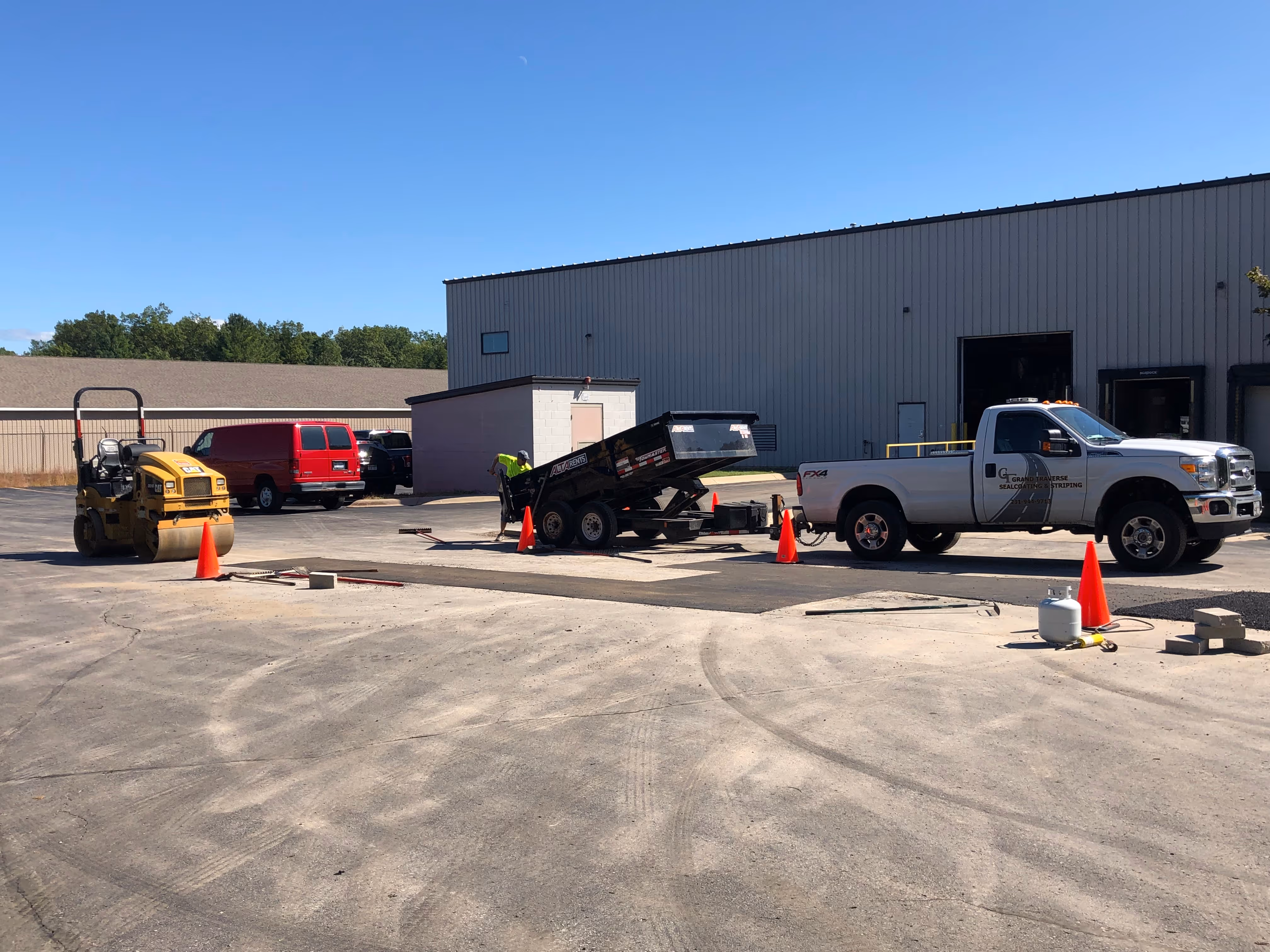 Professional asphalt repair in progress at VP Demand Creations on Aero-Park Dr in Traverse City, MI. A large Caterpillar asphalt roller compacts new patches while a worker in a safety green shirt shovels hot-mix from a raised dump trailer into a prepped repair area secured by orange cones.