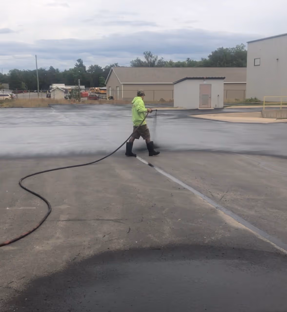 A technician spraying industrial-grade asphalt sealer at the VP Demand Creations parking lot. The sealant is applied over asphalt patches that have undergone a full 30-day curing period, ensuring maximum chemical bonding and surface protection.