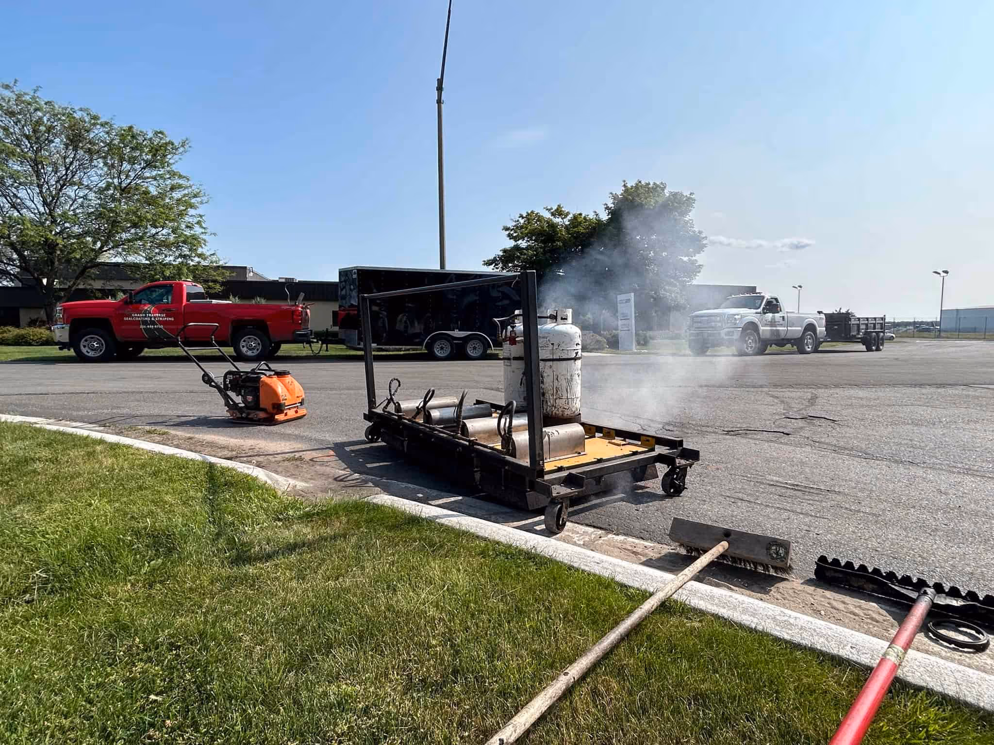Active infrared asphalt repair at the entrance of Century Sun Metal Treating on Aero-Park Dr in Traverse City, MI. The image captures the infrared heating process (visible smoke) used for seamless pavement restoration, featuring a plate compactor and lutes ready for the finish, with two Grand Traverse Sealcoating & Striping service trucks in the background.