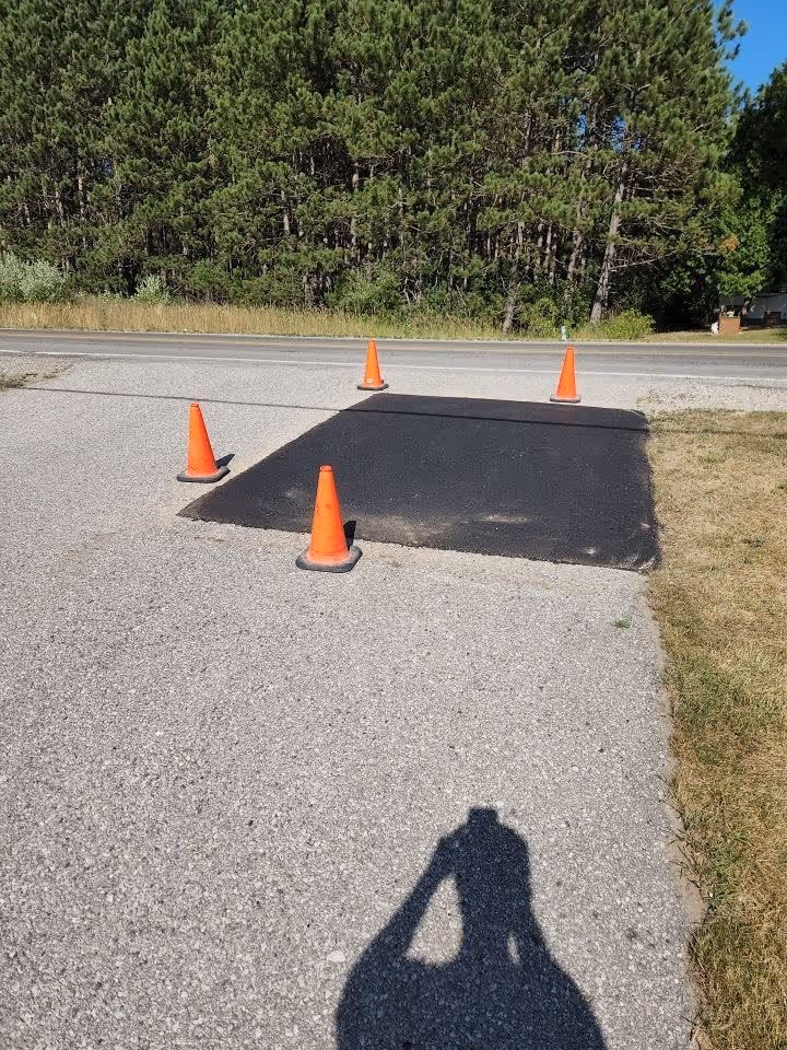A large, professionally compacted asphalt repair patch on a residential driveway located on Bass Lake Rd near Dyer Lake in Traverse City, MI. The image showcases the completed structural restoration secured with orange safety cones to ensure proper curing of the new pavement.