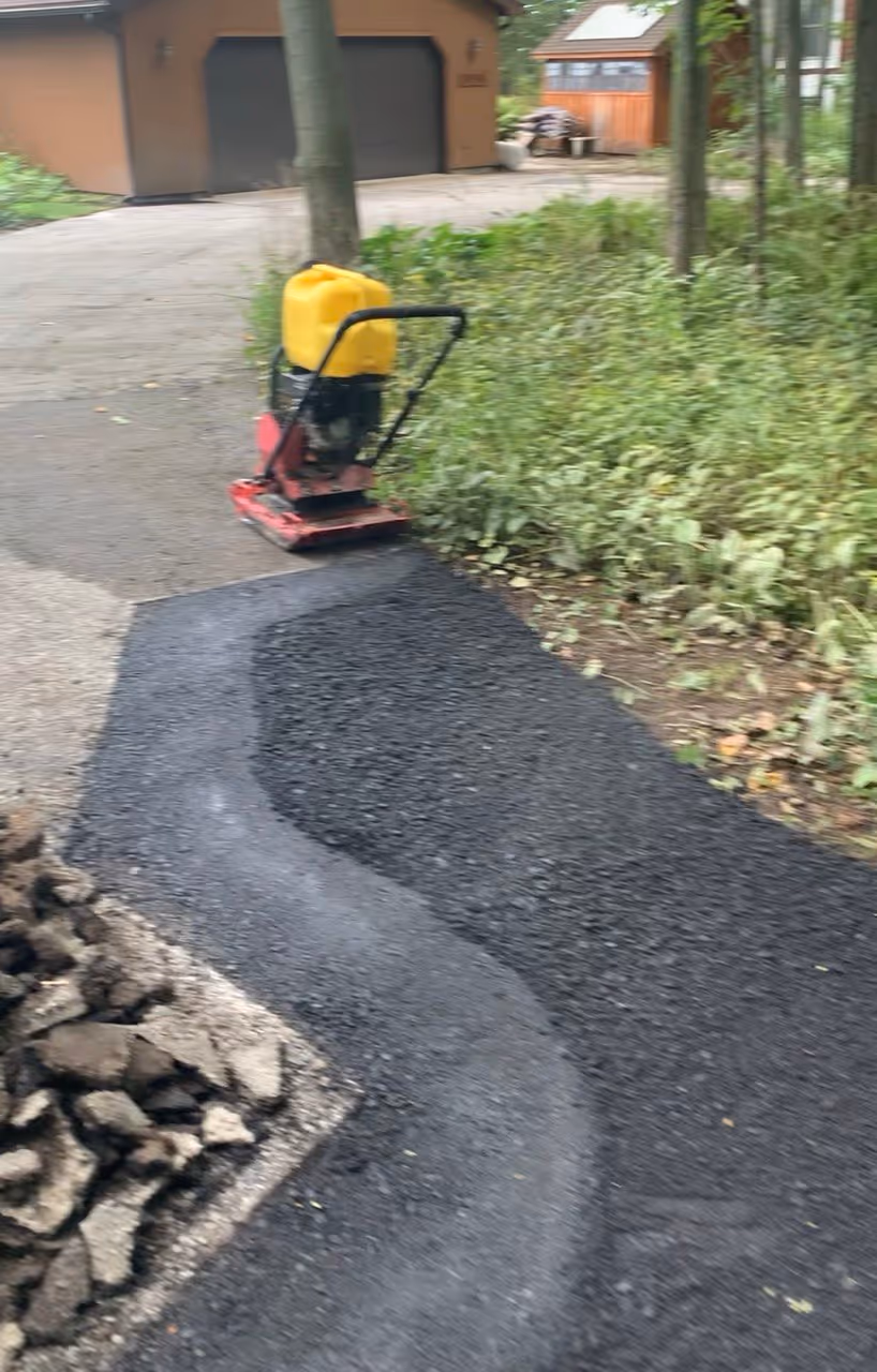 A residential driveway asphalt repair on Shaw Rd in Williamsburg, MI, located off the US-31 corridor. The image shows a stack of removed debris alongside new hot-mix asphalt being stabilized with a specialized plate compactor featuring an integrated water tank for a smooth, professional, and high-durability finish.