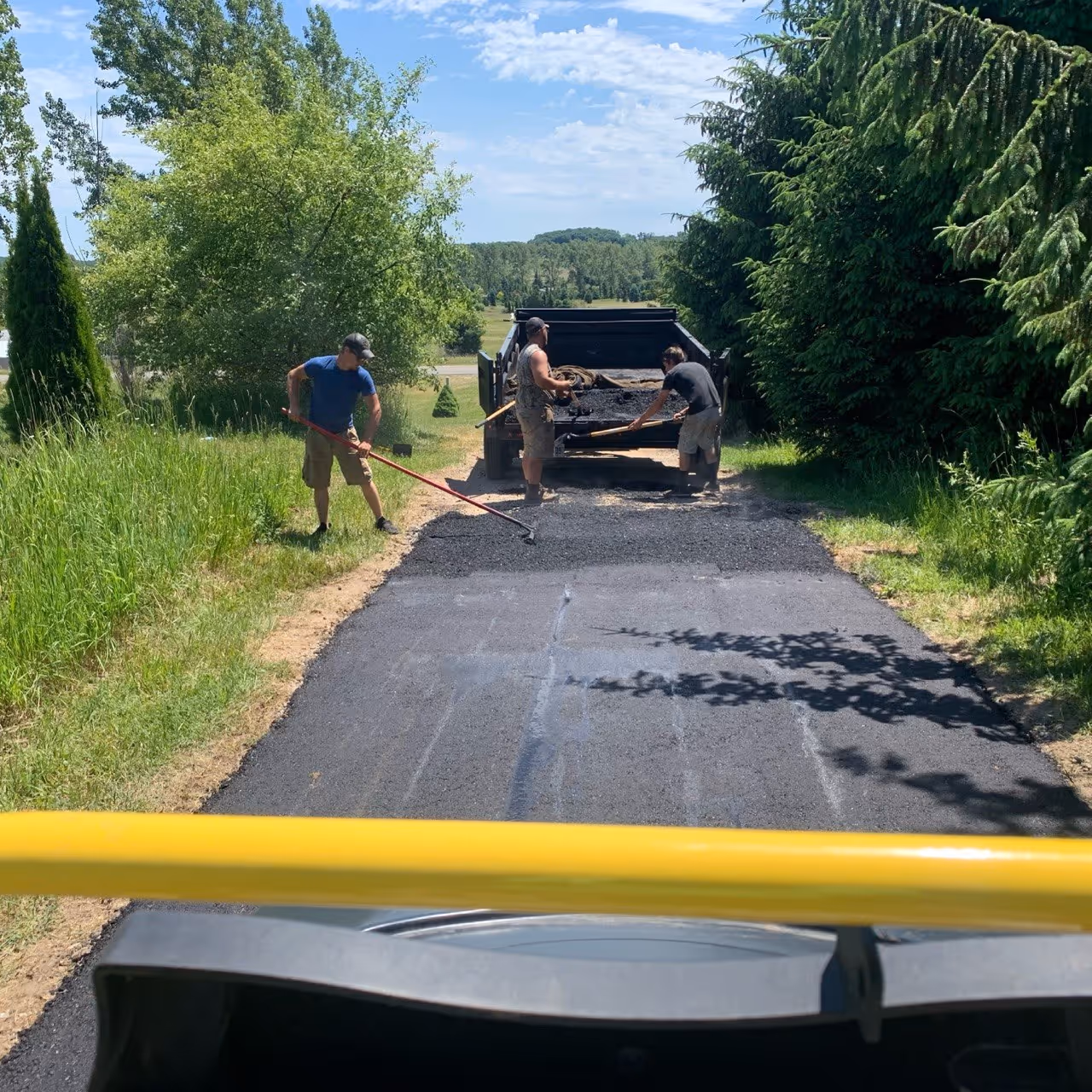 A POV shot from a Bomag roller during a large asphalt driveway repair on E Harry’s Rd in Traverse City, MI. The image shows a professional crew installing steaming hot-mix asphalt, with workers shoveling from a raised dump trailer and luting the surface for a smooth finish across from Breeze Hill Greenhouse and the Great Lakes Humane Society.