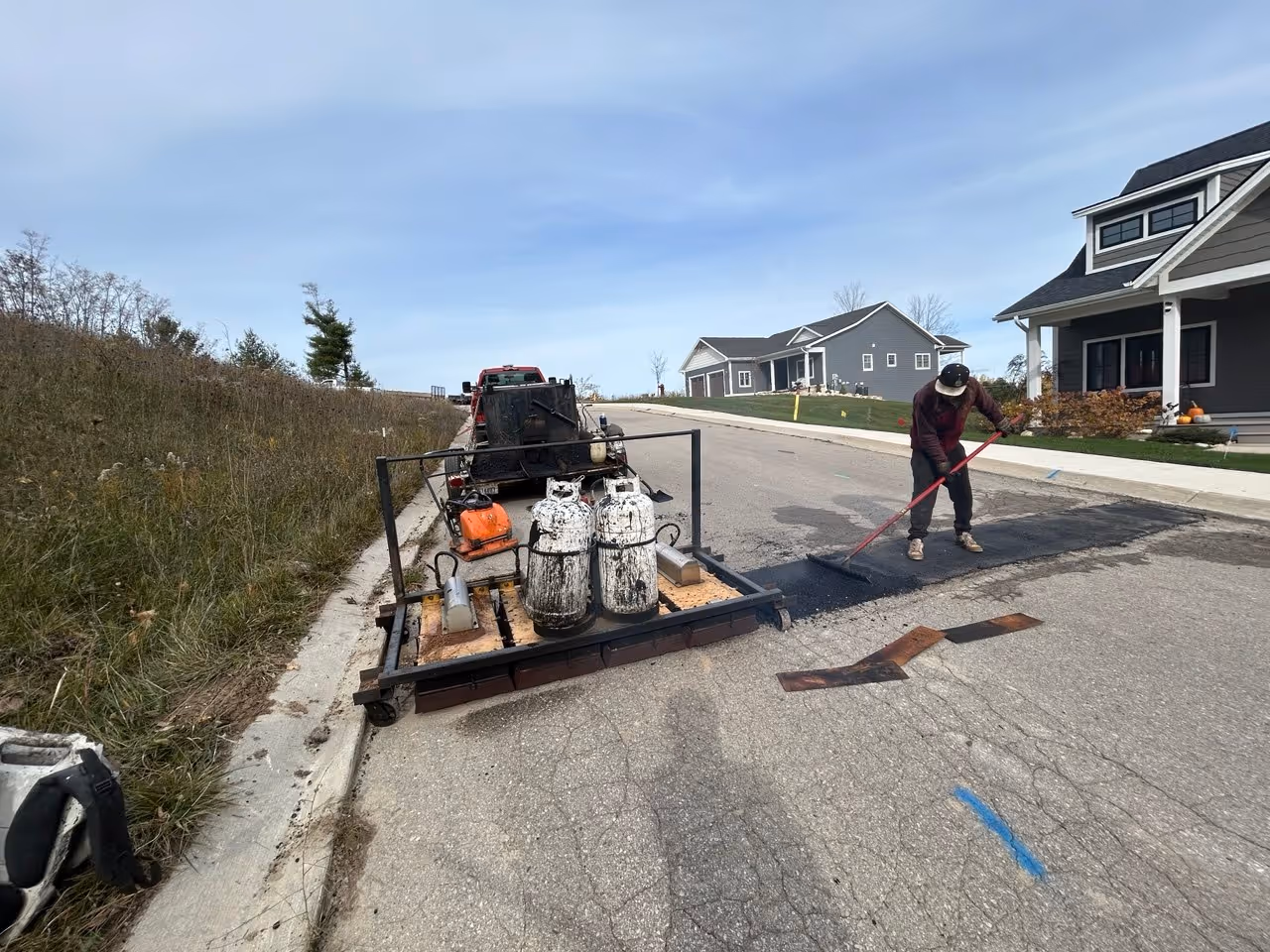 Active infrared asphalt repair fixing a separated paving seam on S Winding Trail in the Morgan Hill neighborhood of Traverse City, MI. The image shows a worker raking steaming, heated asphalt with an asphalt hotbox trailer in the background, creating a seamless thermal bond near the E Traverse Hwy (M-72) corridor.