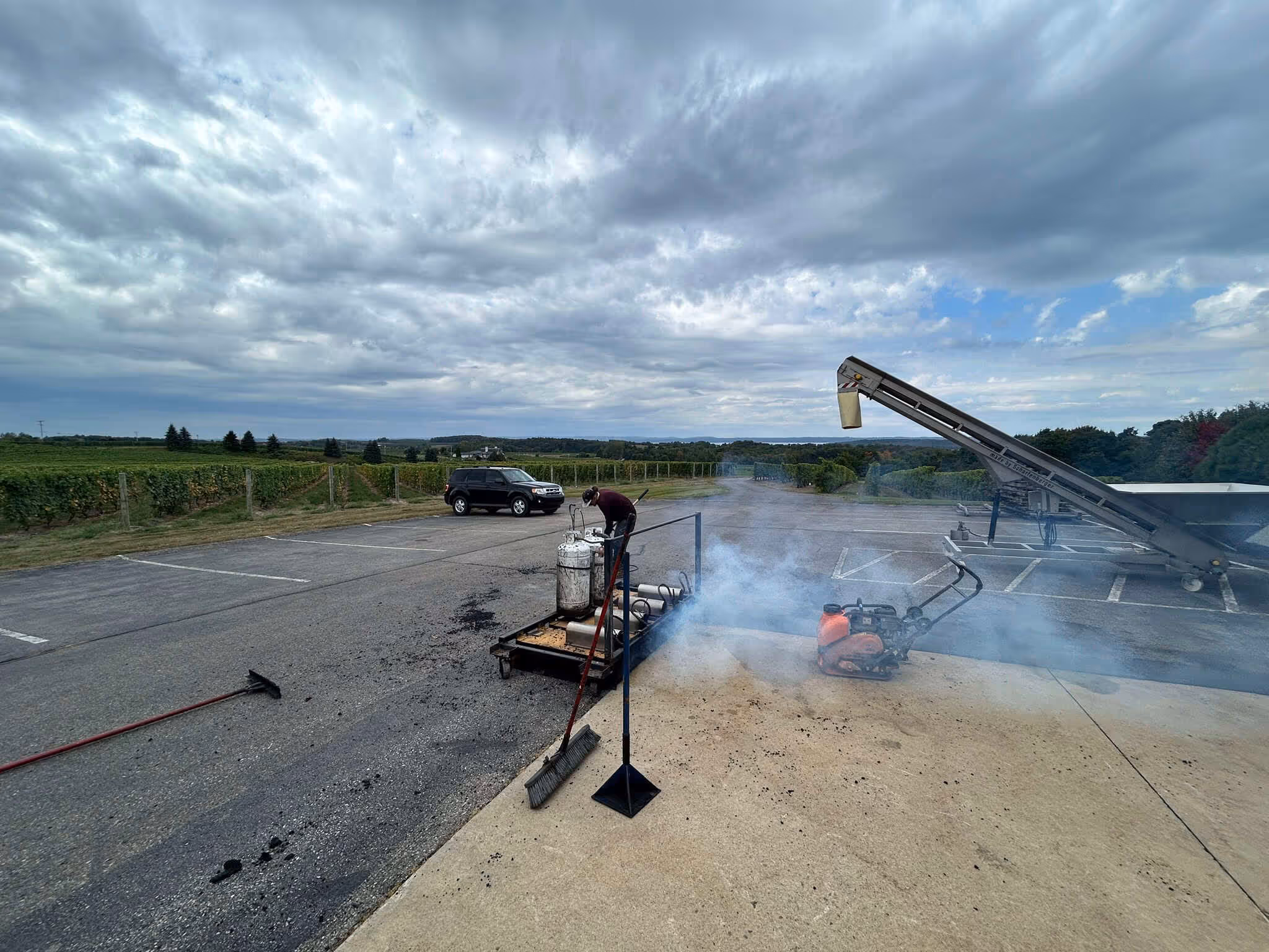 Active infrared asphalt repair at Chateau Grand Traverse Winery on Center Rd, Old Mission Peninsula. The image shows the infrared unit heating the pavement with rising steam, a worker raking the hot-mix asphalt, and a plate compactor prepared to finalize the seamless thermal bond near the Center Road Scenic Overlook.